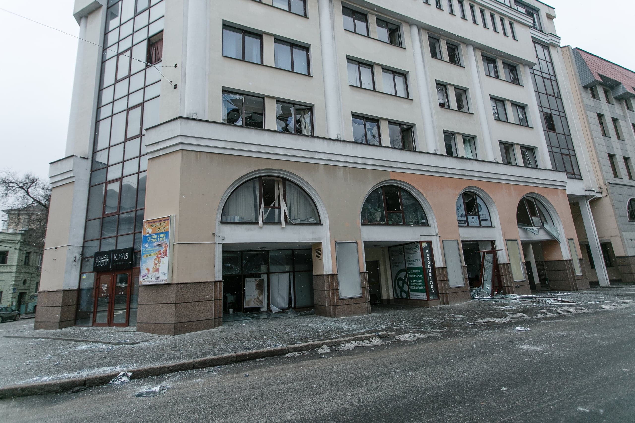 Поврежденное многоэтажное здание с разбитыми окнами на всех этажах. Осколки стекла покрывают тротуар. На фасаде видны вывески "KRASS GROUP", "K PAS" и "АВТОШКОЛА".