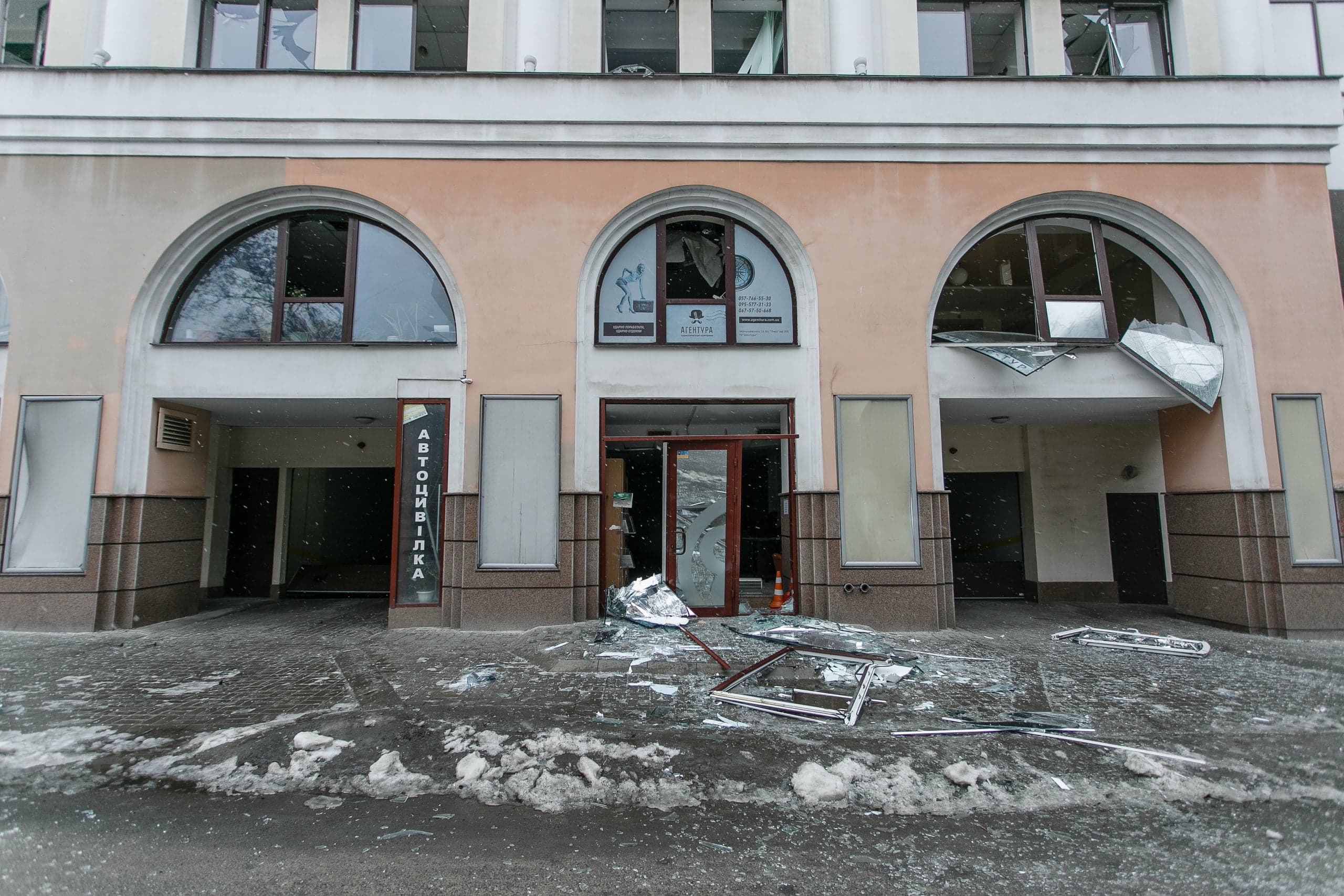 Поврежденный фасад здания с выбитыми окнами и дверями. Осколки стекла и обломки лежат на тротуаре. На вертикальной вывеске "АВТОМИЙКА".