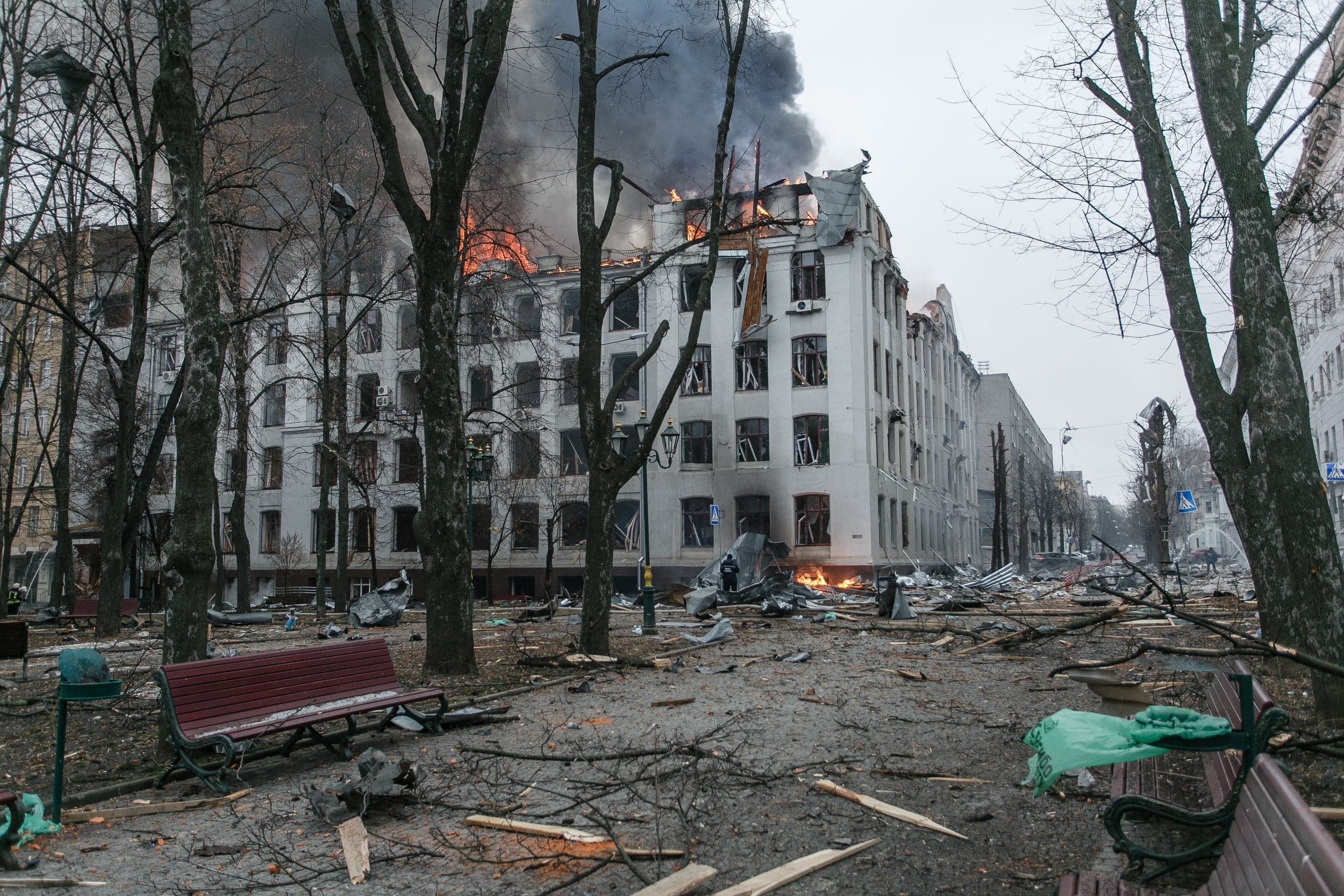 Разрушенное здание в огне с густым дымом, на переднем плане заваленный обломками парк с голыми деревьями и скамейками.