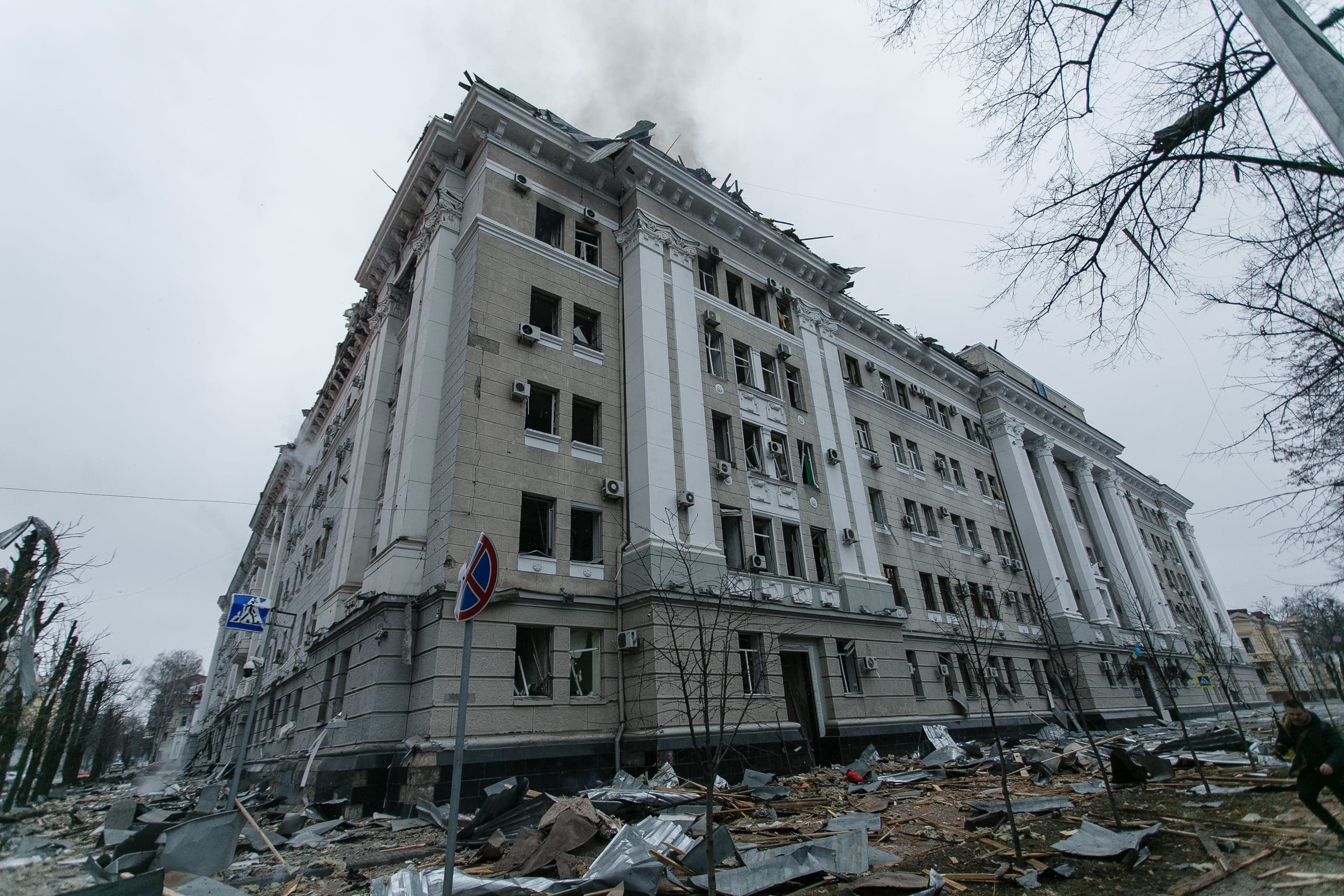 Сильно поврежденное многоэтажное здание с выбитыми окнами, дымом на крыше и обломками на улице.