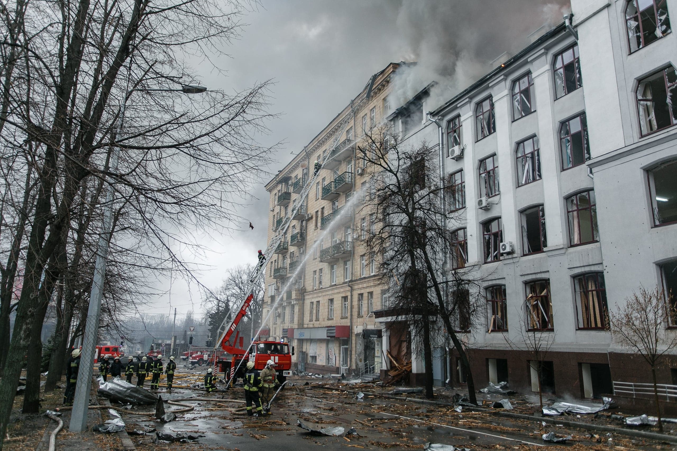 Пожарные тушат огонь на верхних этажах поврежденного здания; улица усыпана обломками.