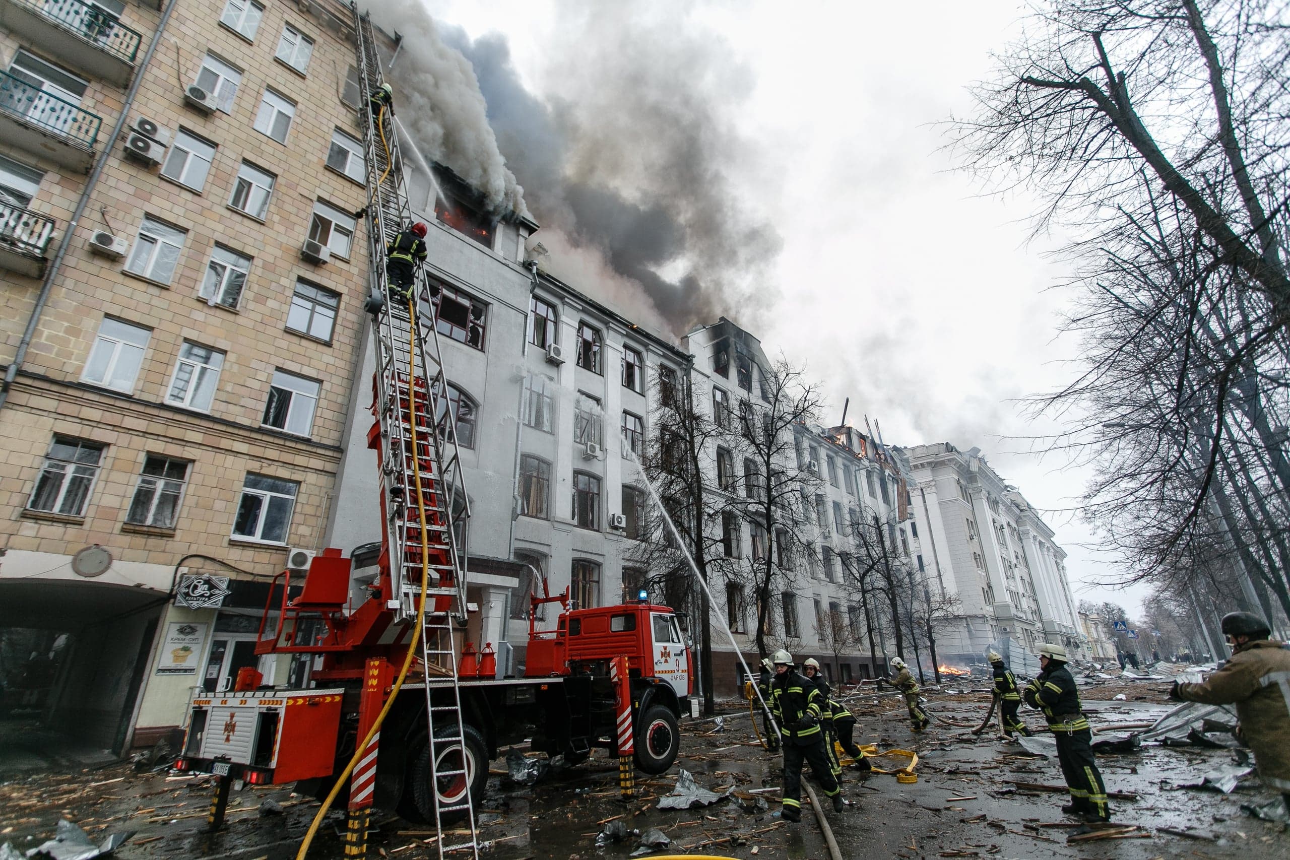 Пожарные тушат горящее многоэтажное здание с помощью автолестницы и брандспойтов; густой дым над крышей. Разрушенная улица.