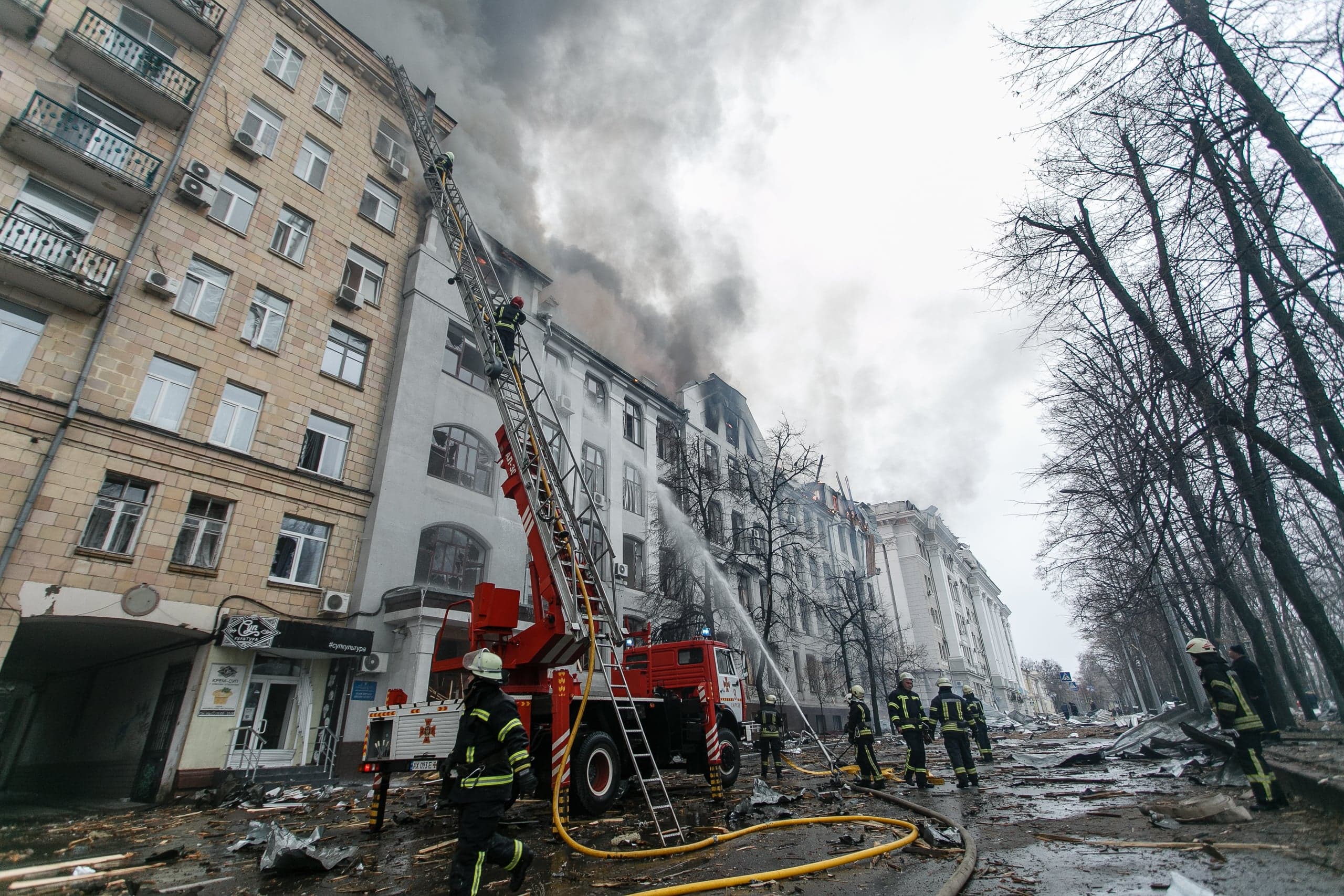 Пожарные тушат сильный пожар в многоэтажном здании с автолестницы. Густой дым валит из верхних этажей; улица завалена обломками.