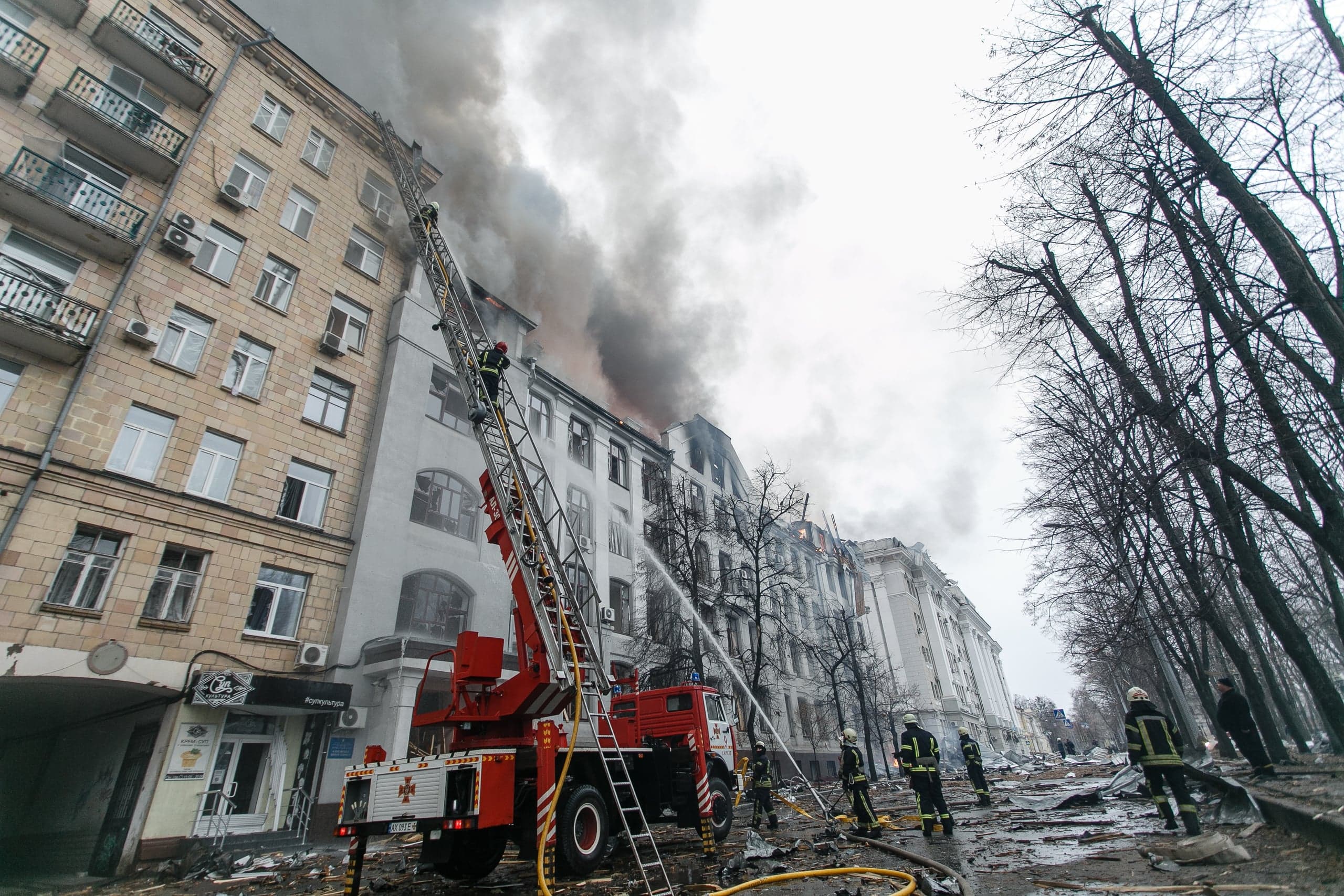 Пожарные тушат сильный пожар в верхних этажах разрушенного городского здания с помощью автолестницы и брандспойтов. Улица завалена обломками.