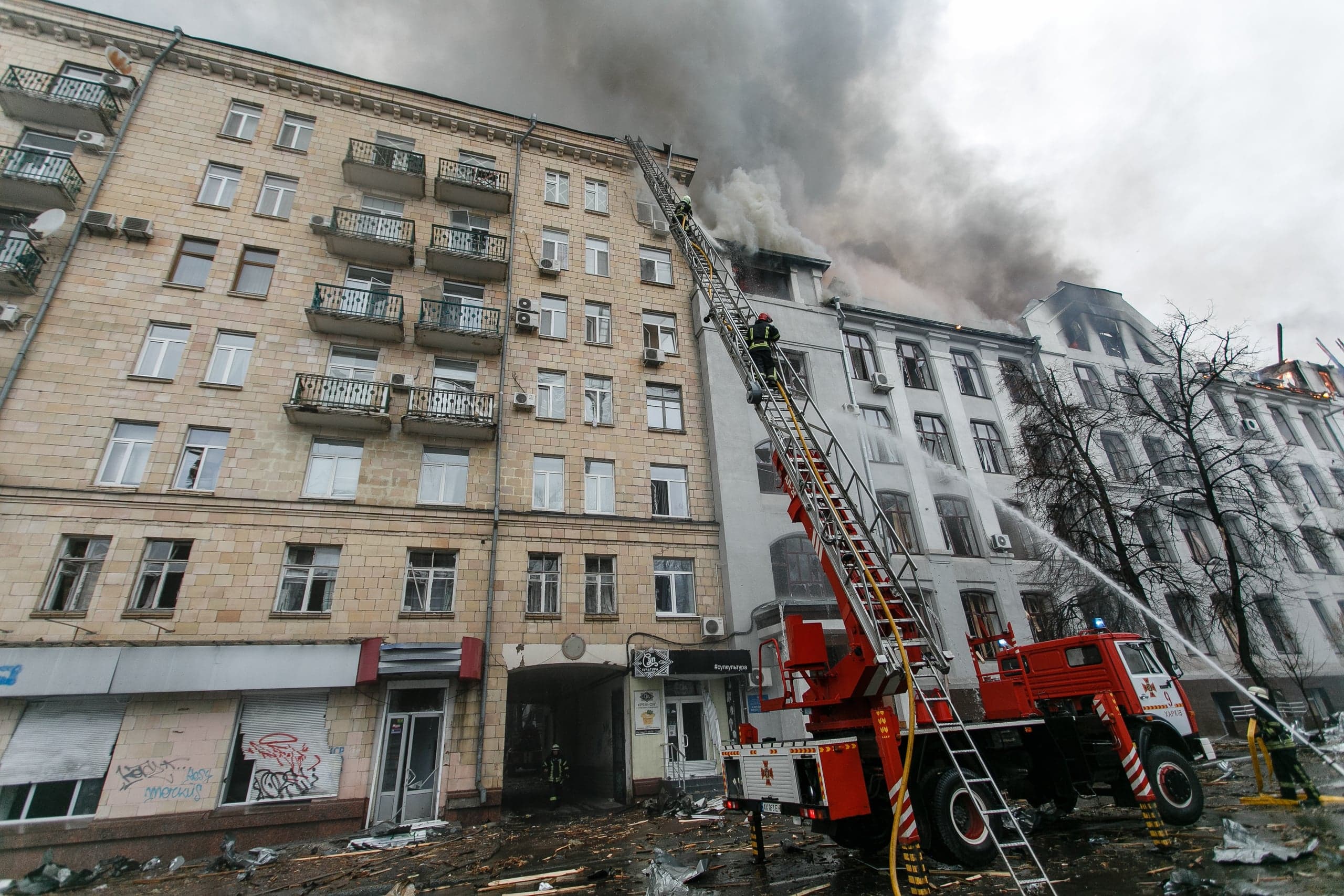 Пожарная машина с выдвижной лестницей и пожарные тушат сильный пожар в верхних этажах многоквартирного дома, охваченного густым дымом.