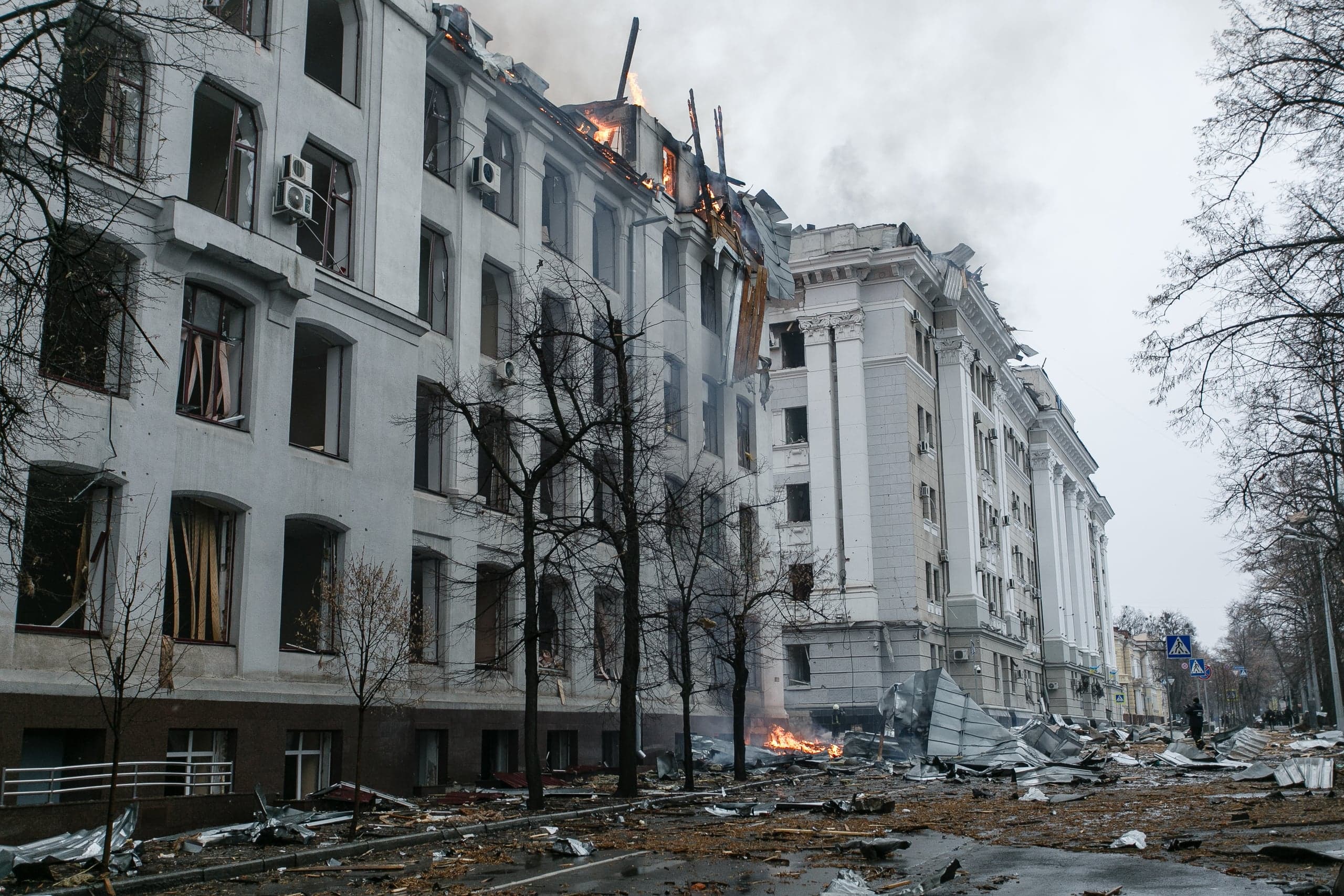 Поврежденное административное здание с горящей крышей. Улица перед фасадом завалена обломками и мусором.