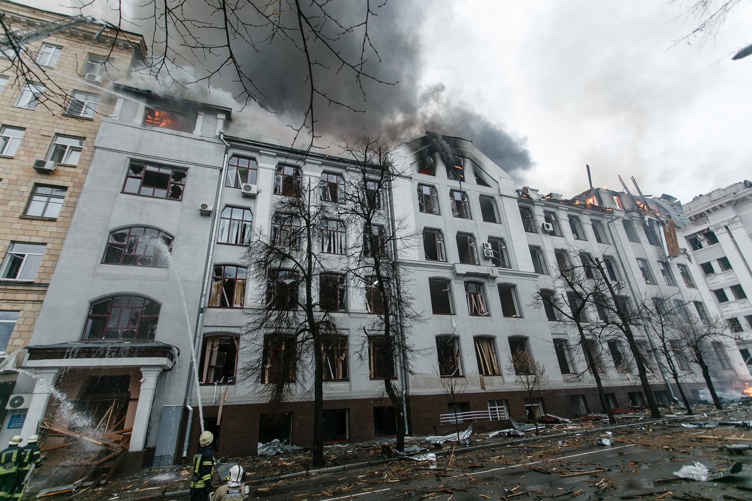 Пожарные тушат сильно поврежденное многоэтажное здание с выбитыми окнами, из которого идет густой черный дым.