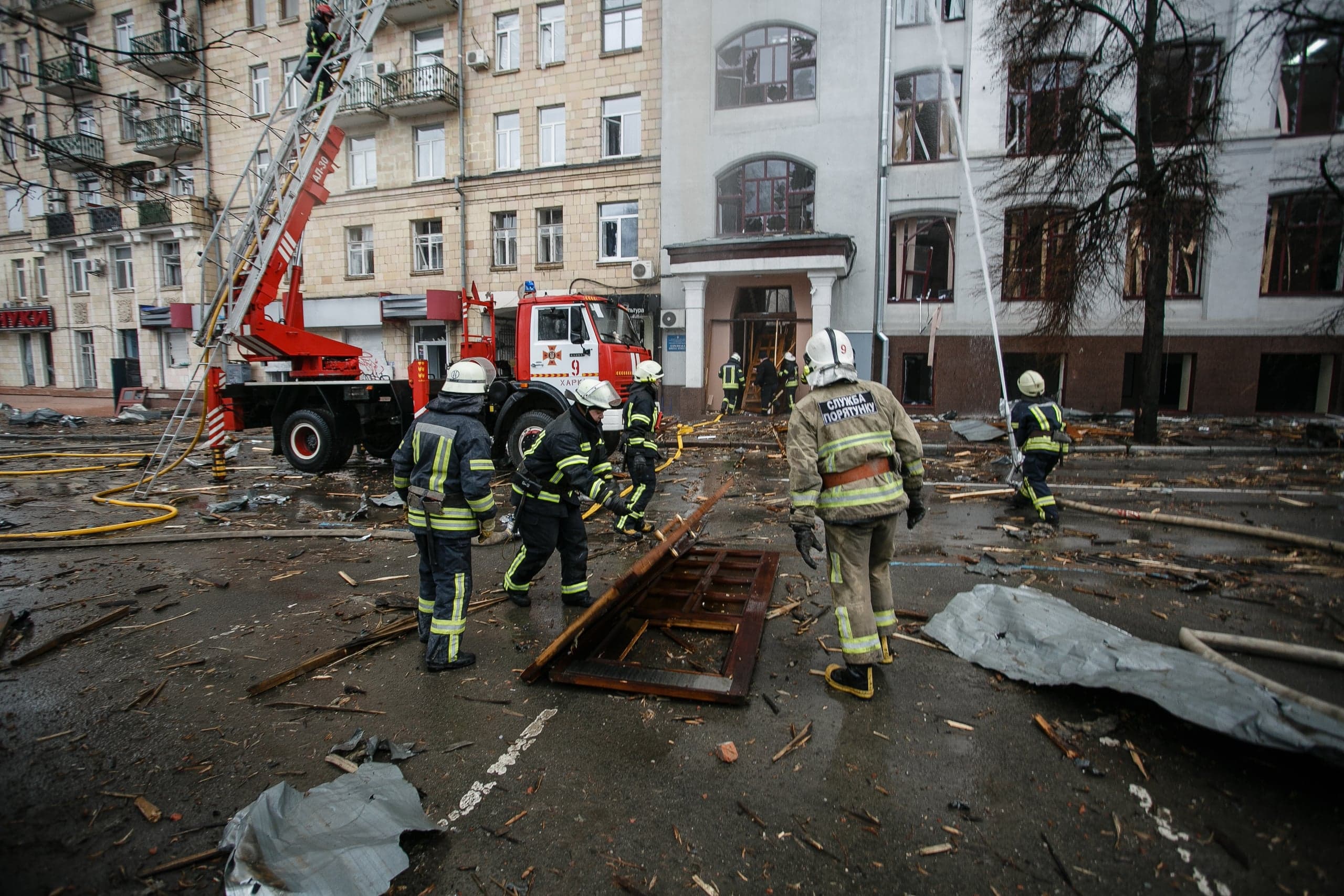 Украинские спасатели работают среди обломков улицы перед поврежденным зданием. Слева пожарная машина с выдвижной лестницей. На форме надпись: "СЛУЖБА ПОРЯТУНКУ".