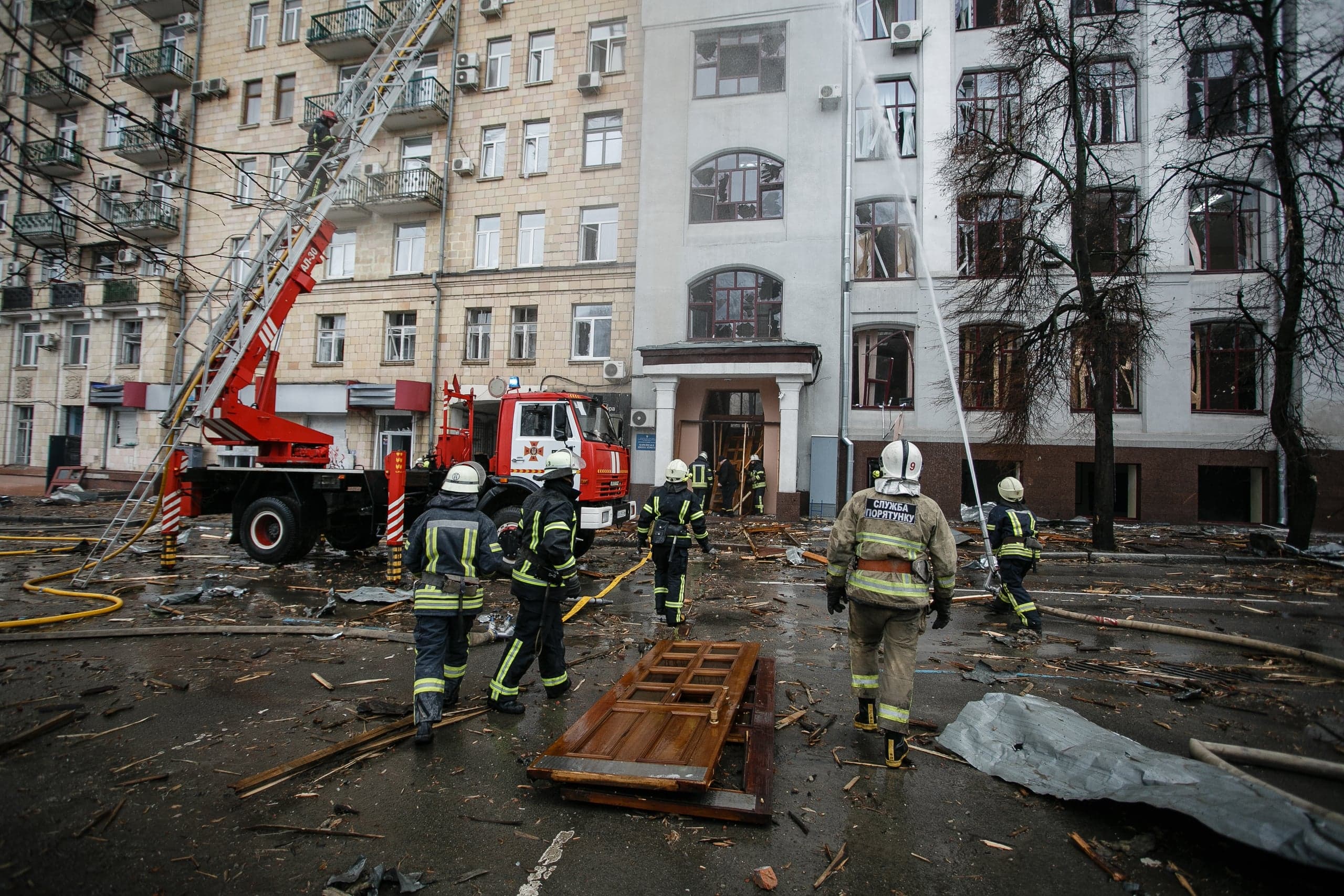 Пожарные и спасатели работают у сильно поврежденного здания, тушат остатки огня. Улица завалена обломками. Виден пожарный автомобиль с выдвинутой лестницей. На форме спасателя надпись: "СЛУЖБА ПОРЯТУНКУ".