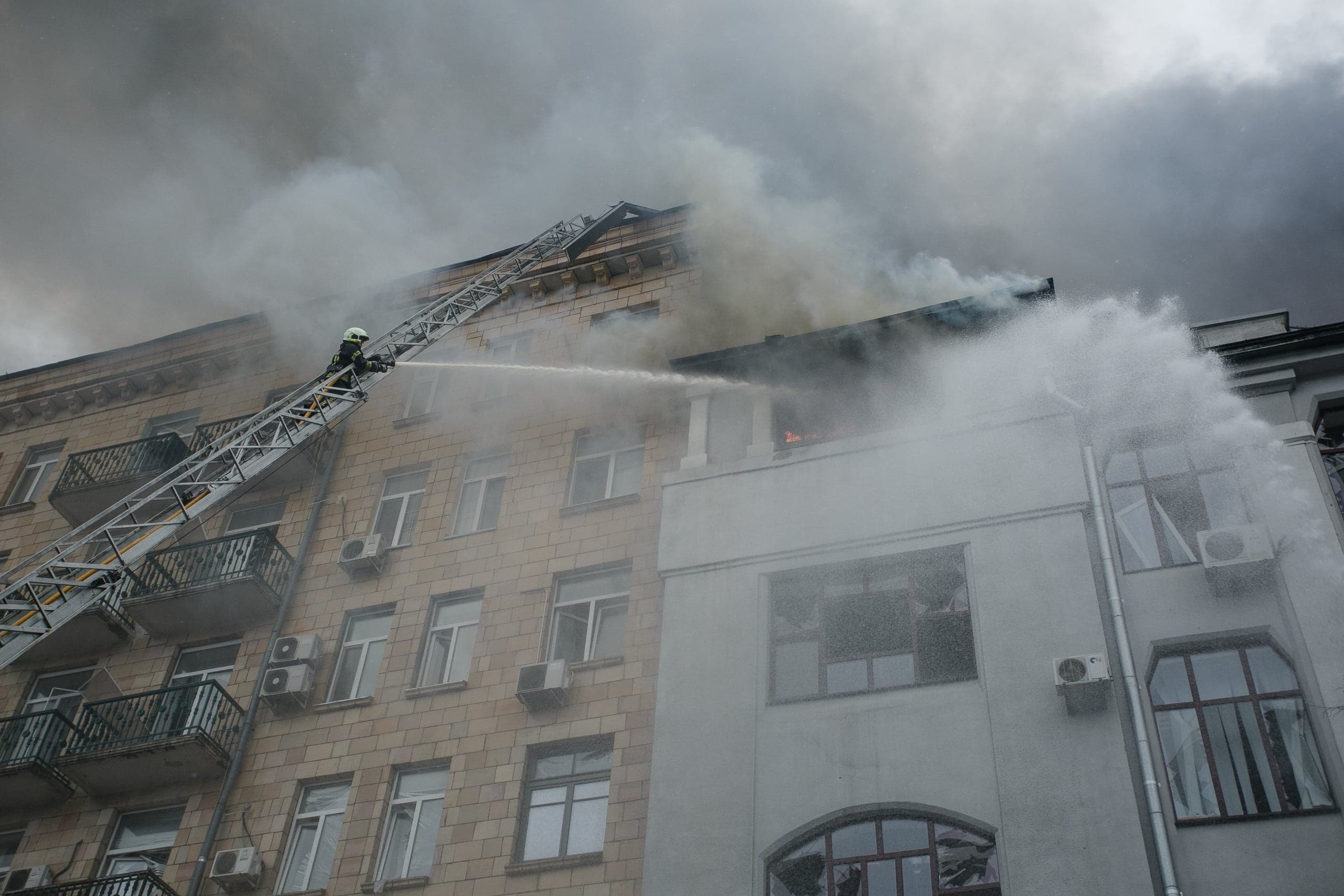Пожарный на выдвижной лестнице тушит пожар на крыше многоэтажного здания, окутанного дымом.