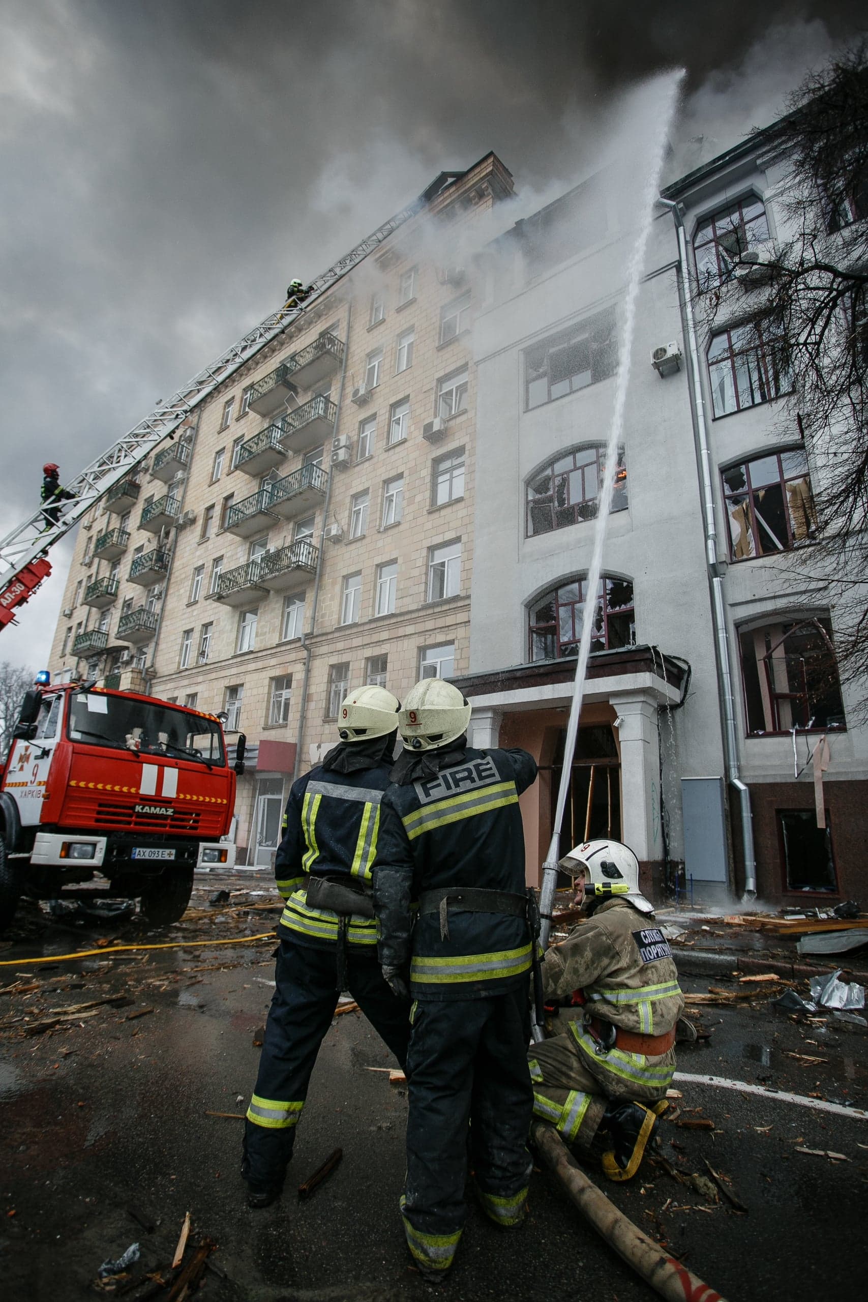 Пожарные тушат пожар в поврежденном многоэтажном здании, используя струи воды и автолестницу. На переднем плане — спасатели; на улице — обломки и пожарный автомобиль "КАМАЗ".