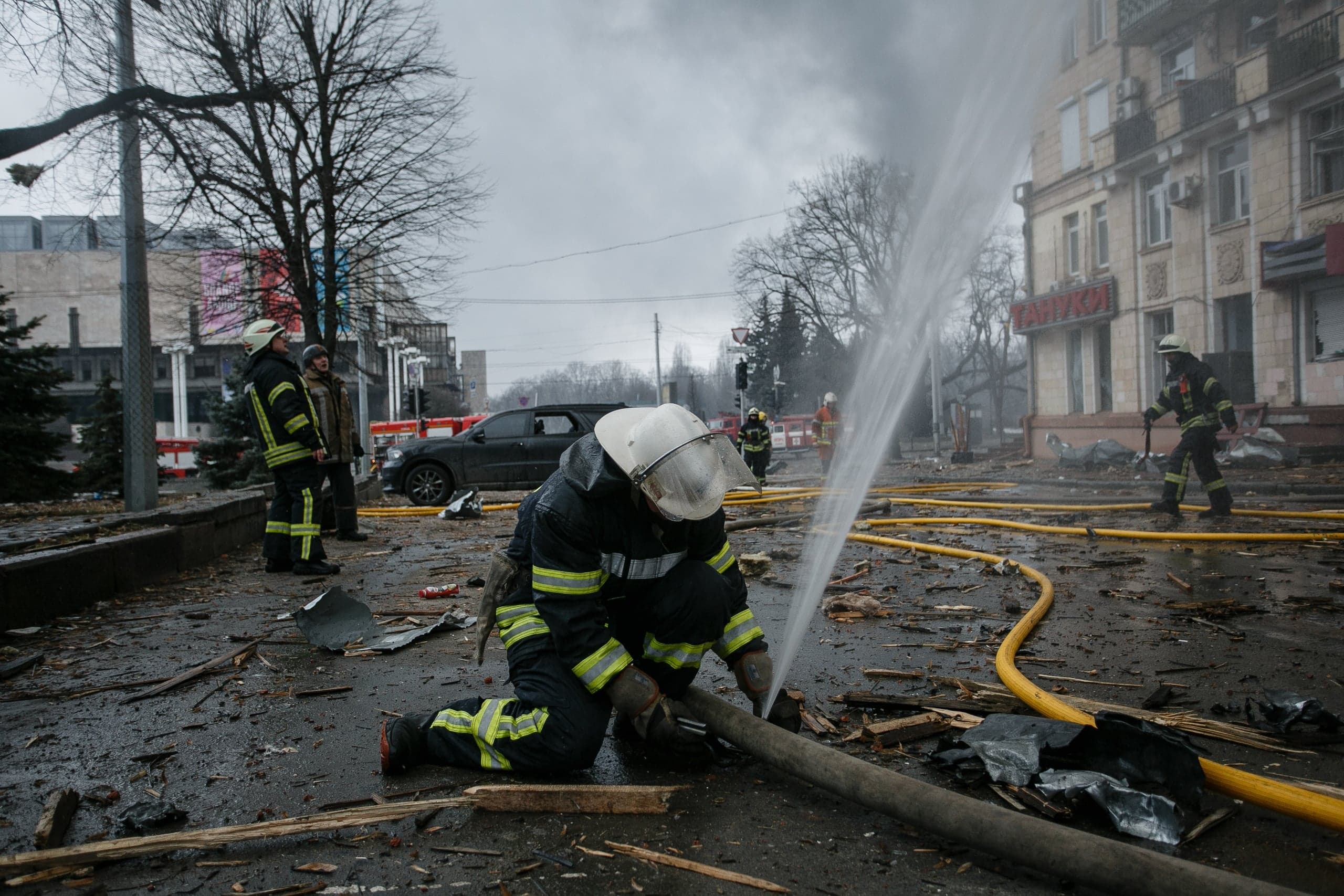 Пожарный на коленях на заваленной обломками городской улице, из шланга бьет мощная струя воды.