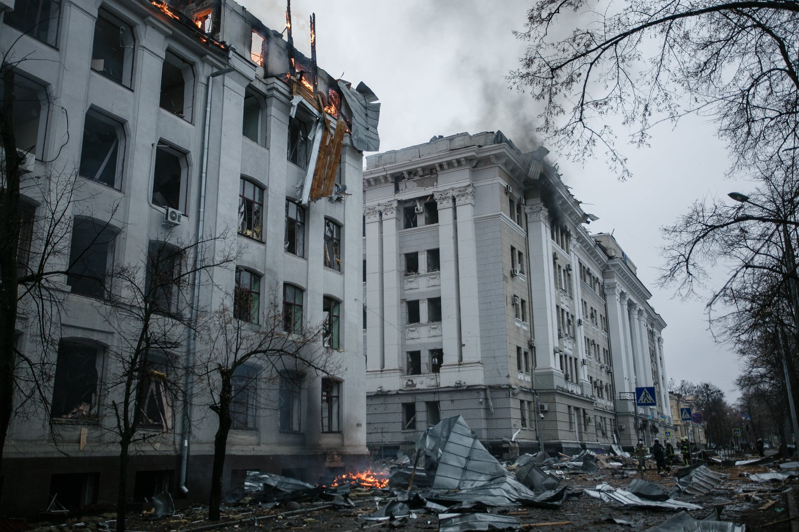 Разрушенные обстрелом административные здания; огонь и дым на крыше, улица усыпана обломками.