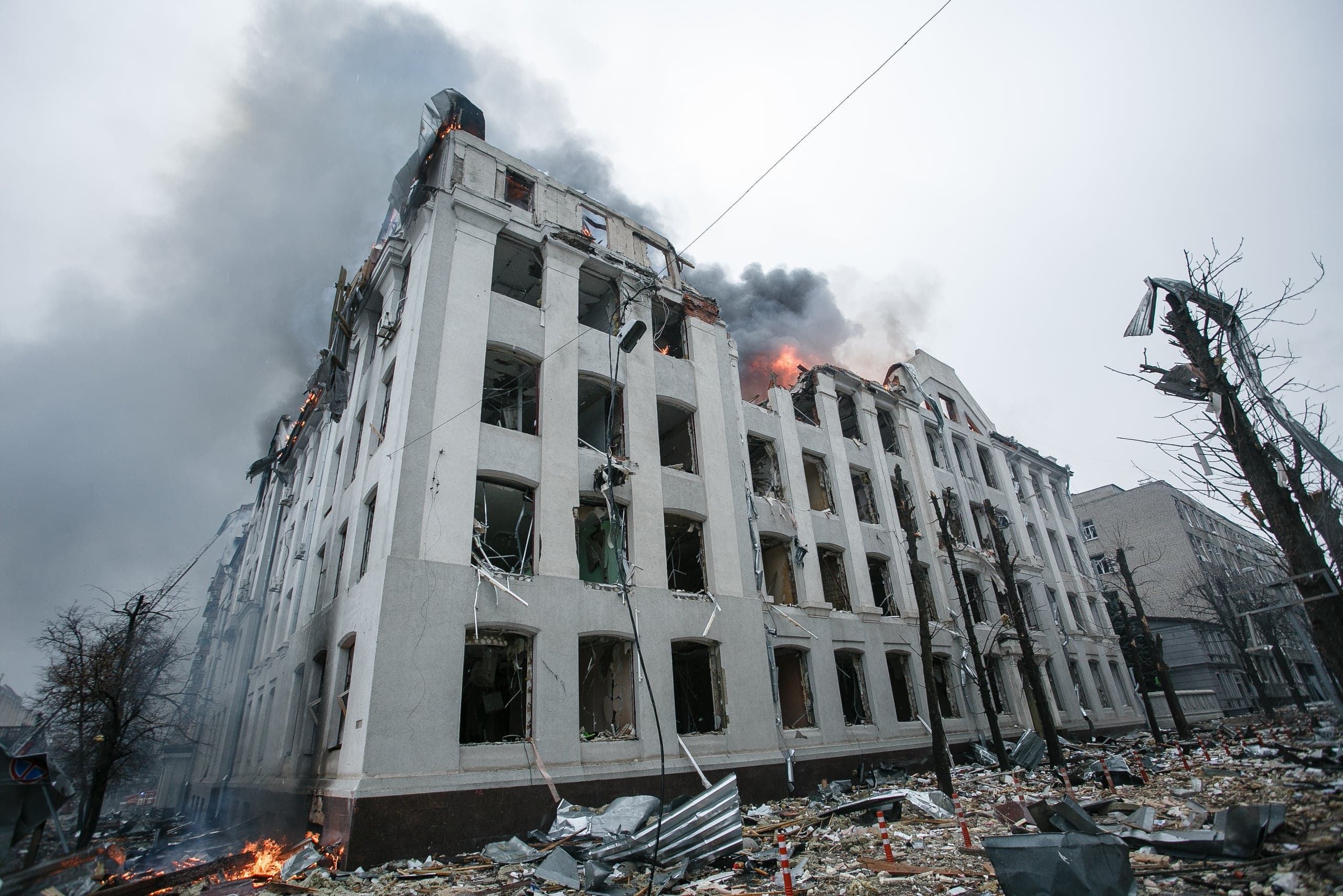 Поврежденное, горящее белое многоэтажное здание с выбитыми окнами и черным дымом на фоне серого неба. Вокруг обломки и мусор.
