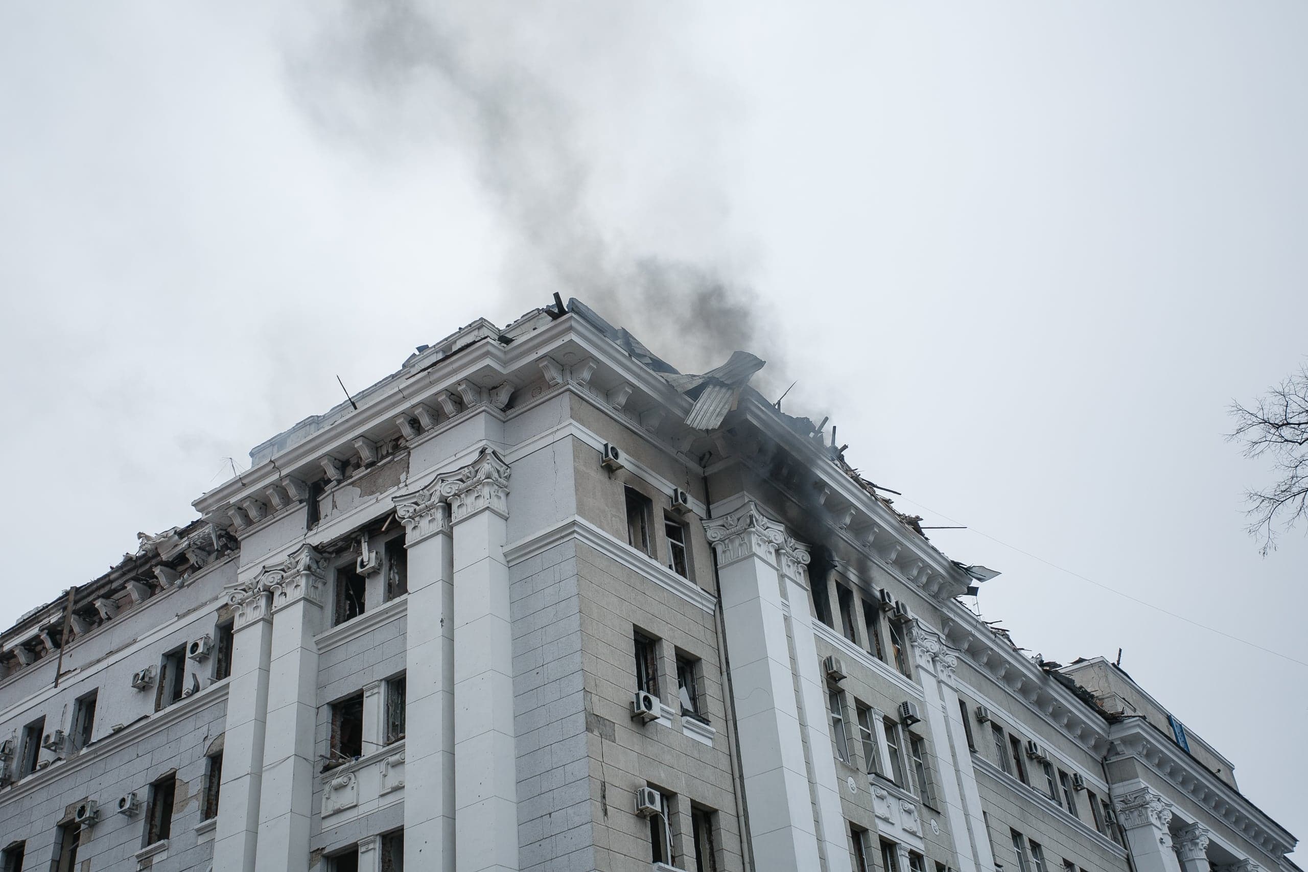 Поврежденное здание в стиле сталинского ампира с разрушенной крышей, из которой поднимается черный дым, на фоне серого неба.