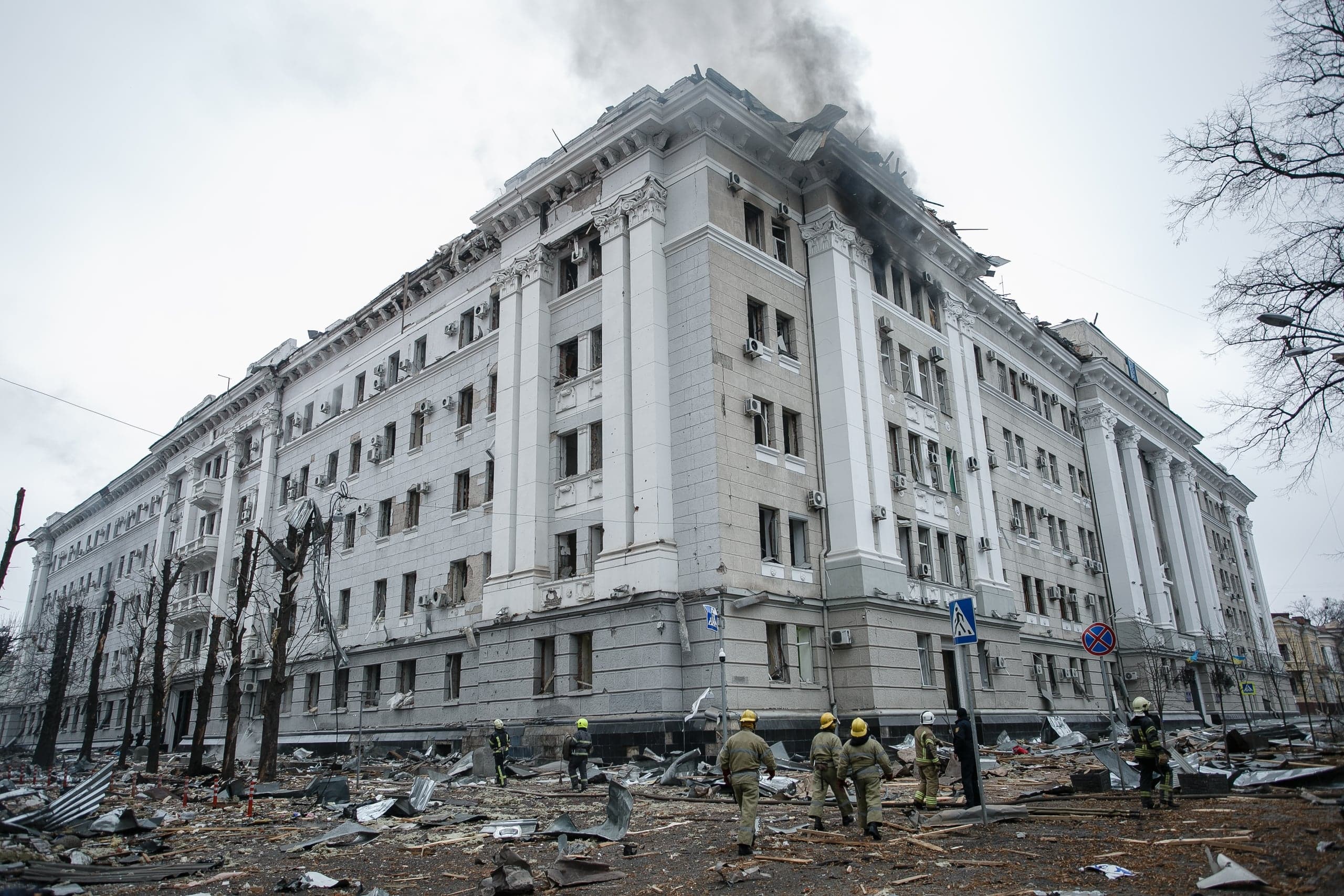 Разрушенное здание областной администрации с дымящейся крышей и выбитыми окнами; спасатели разбирают завалы на площади перед ним.