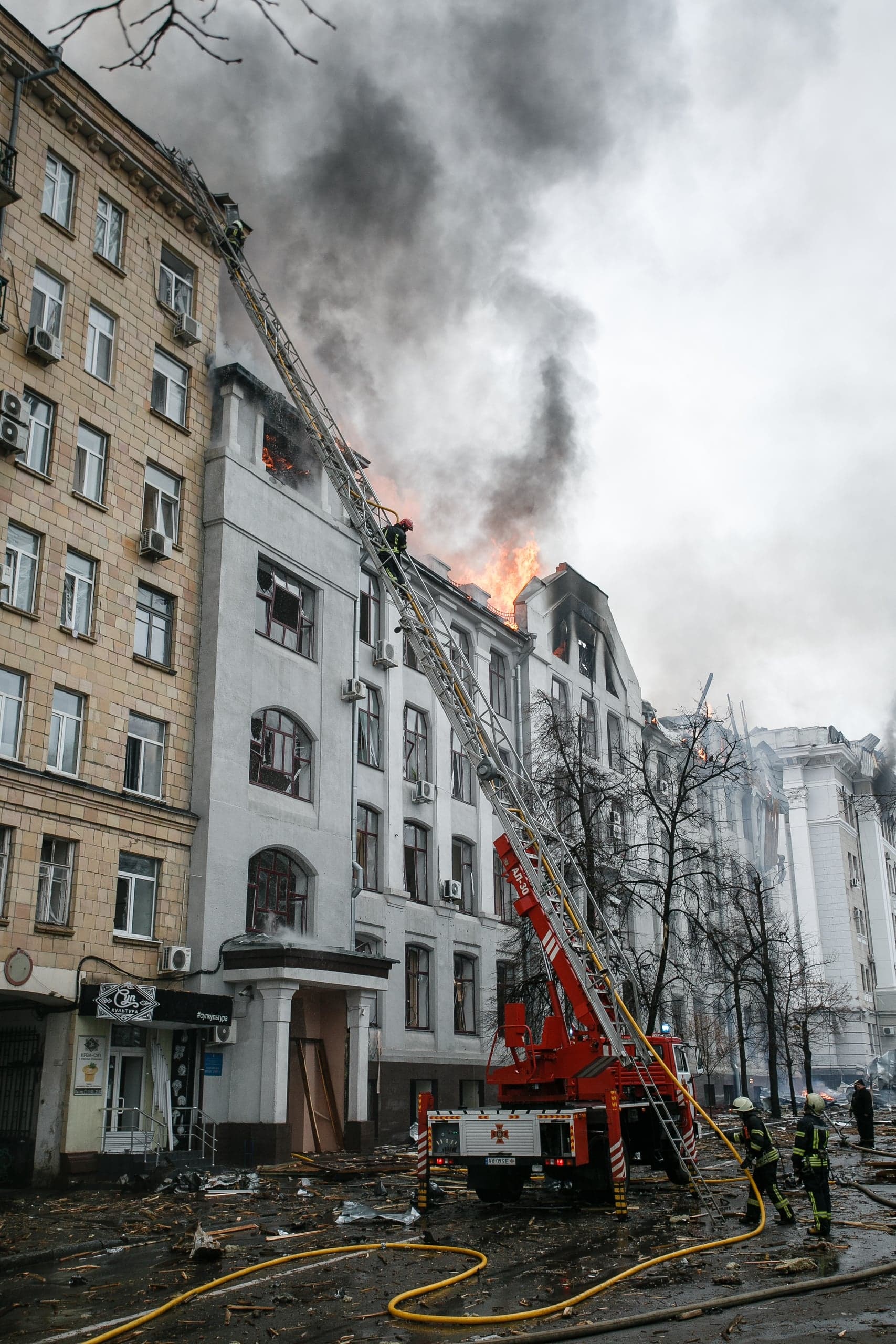 Пожарные тушат сильный пожар в верхних этажах поврежденного взрывом многоэтажного здания. На улице обломки и пожарная техника с выдвижной лестницей.