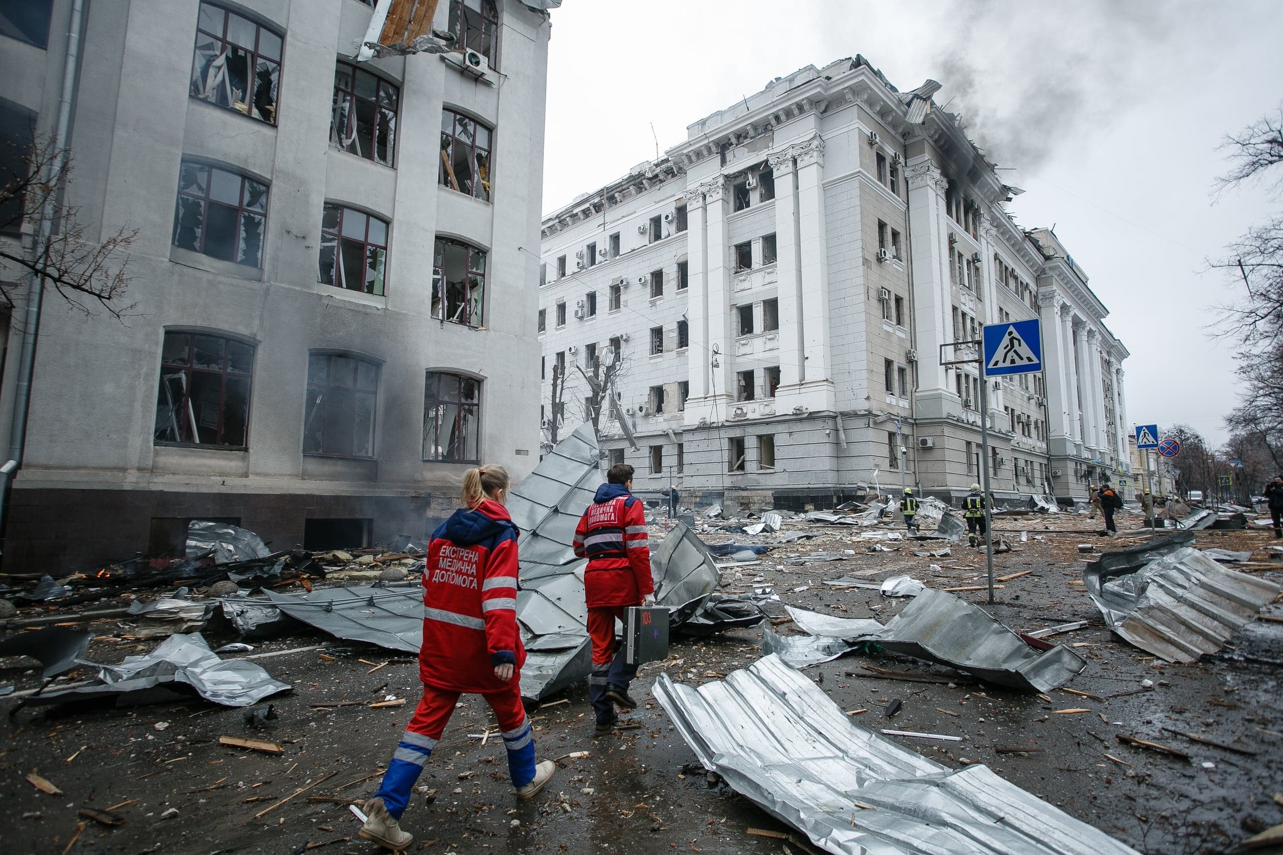 Двое парамедиков в форме "ЕКСТРЕНА МЕДИЧНА ДОПОМОГА" идут по улице, заваленной обломками, мимо сильно поврежденных зданий.