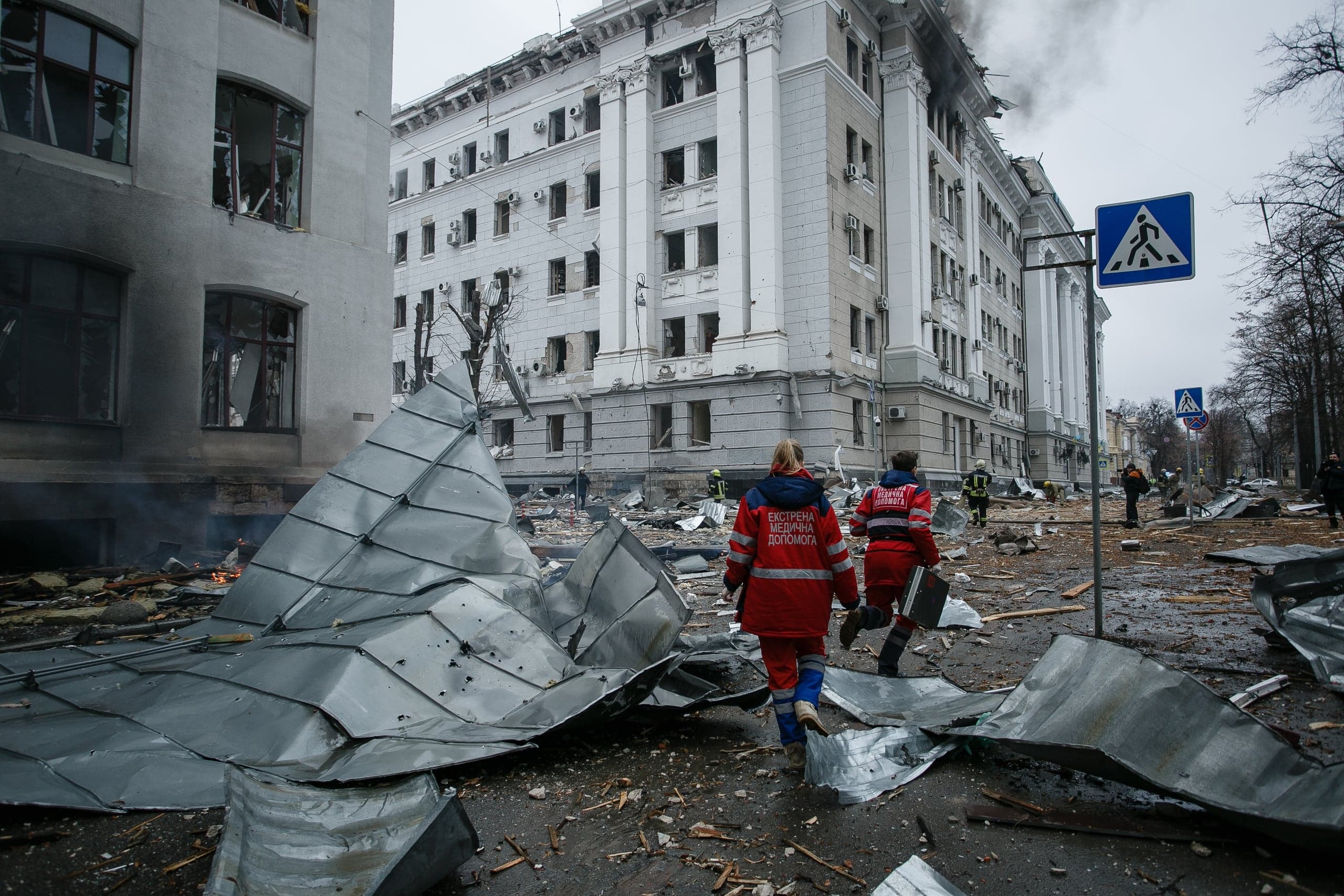 Медики "ЕКСТРЕНА МЕДИЧНА ДОПОМОГА" проходят по разрушенной улице, усеянной обломками и помятым металлом. На заднем плане сильно поврежденное взрывом административное здание, из которого идет дым.