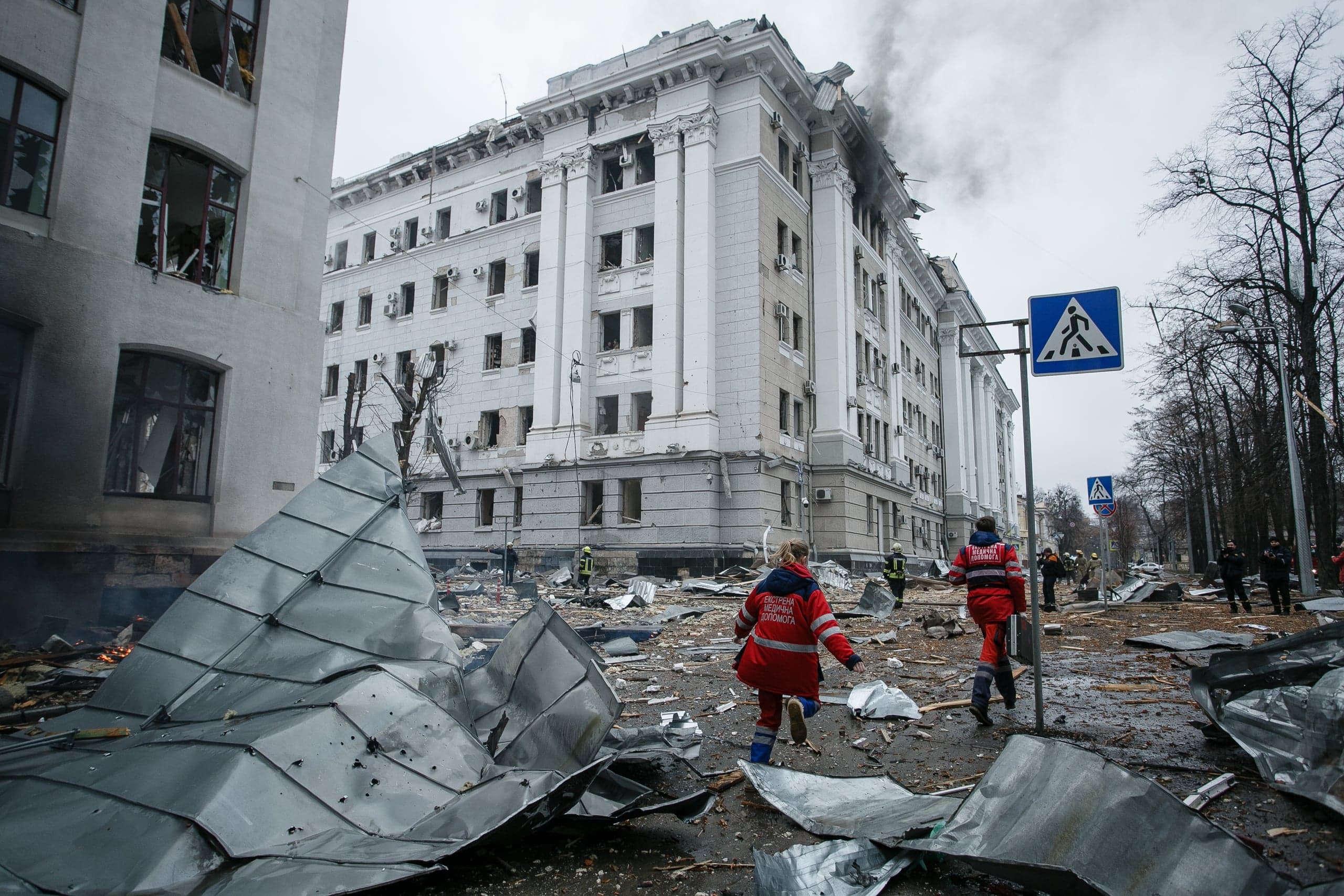 Разрушенное административное здание с дымом на фоне. Улица завалена обломками крыши; спасатели в красной форме идут среди руин.