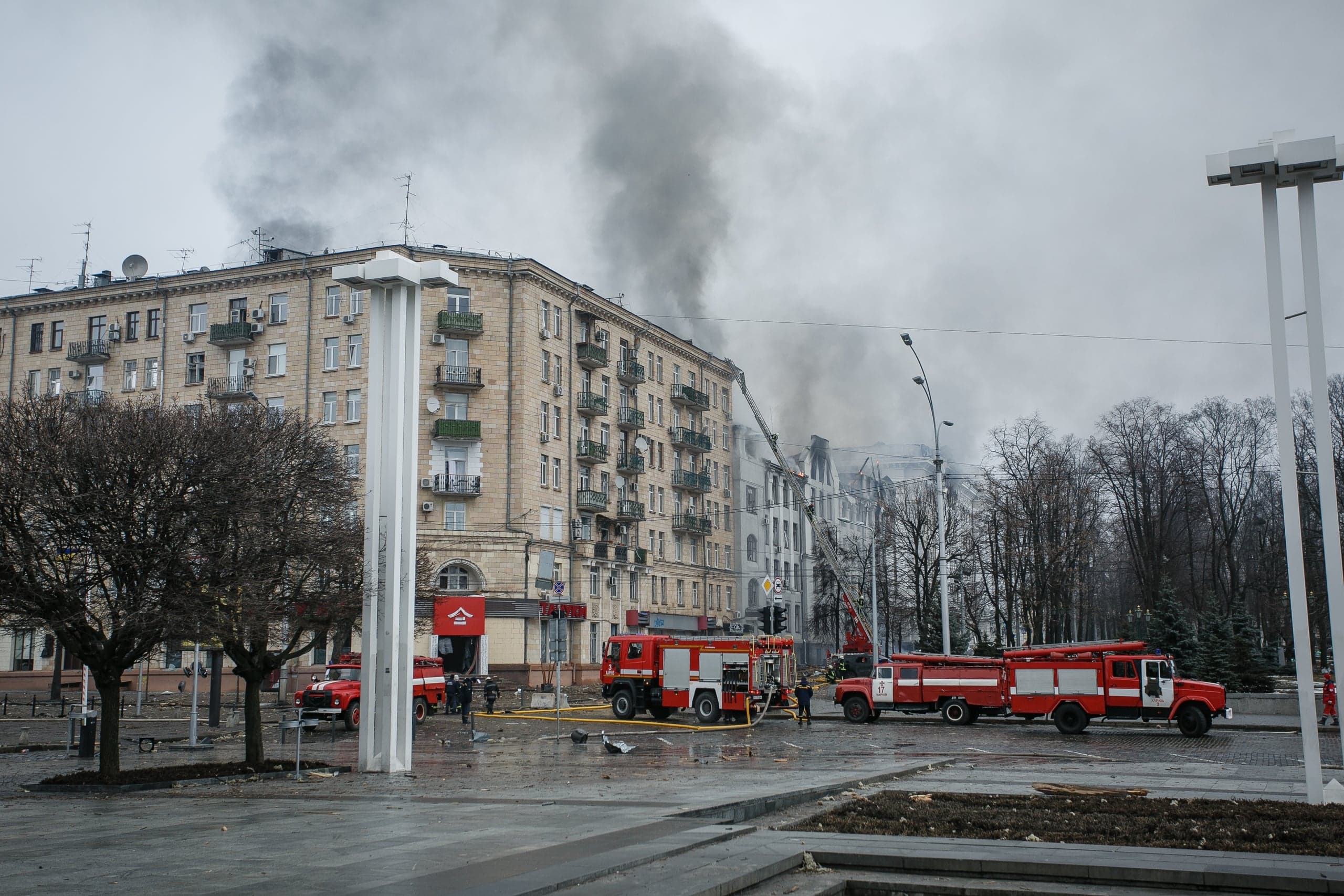 Пожар в многоэтажном здании: густой серый дым над крышей, на площади работают пожарные машины и спасатели.