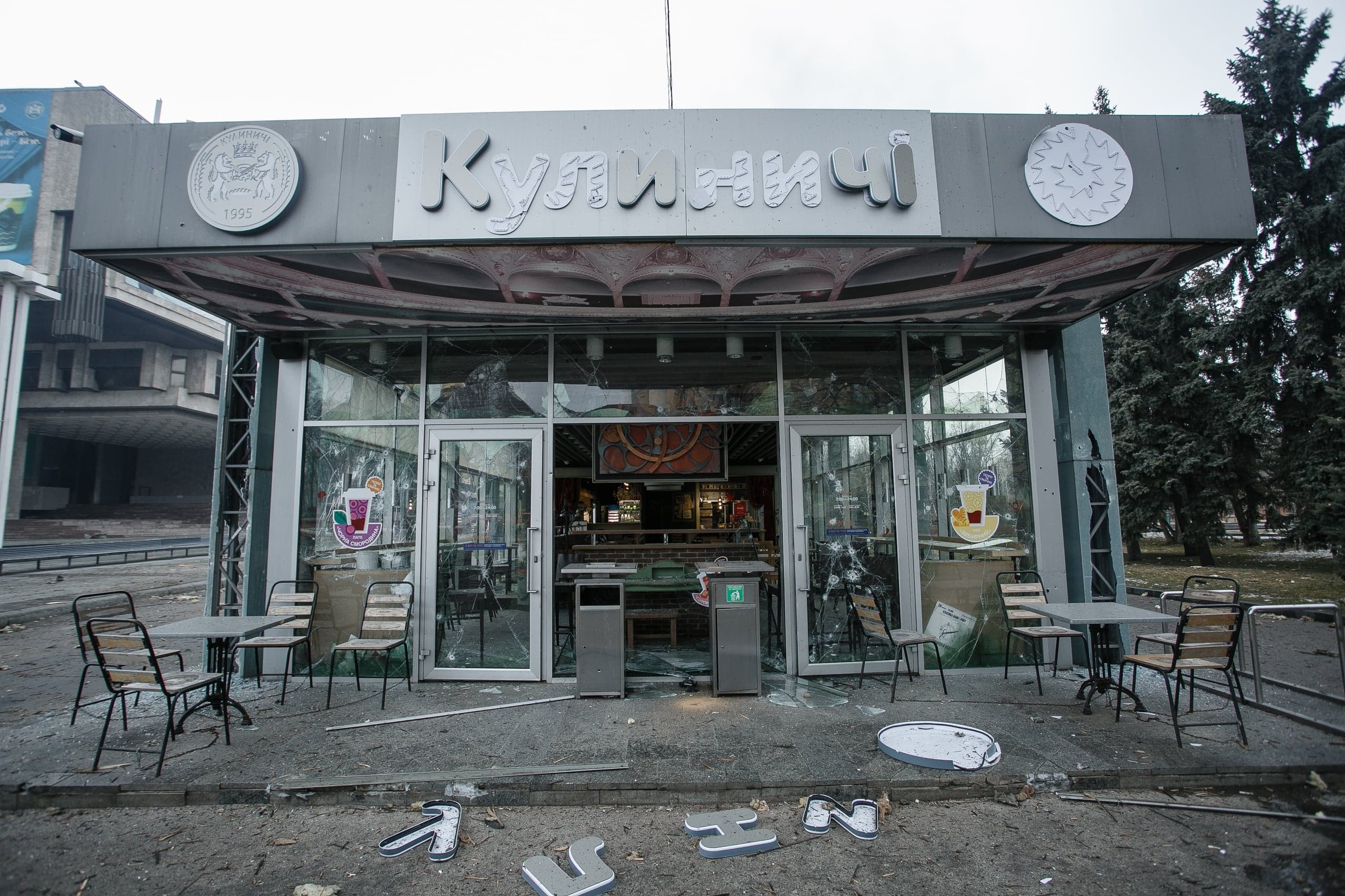 Сильно поврежденное кафе "Кулиничі" с разбитыми окнами и дверями. На земле перед входом лежат осколки стекла и отвалившиеся буквы вывески.