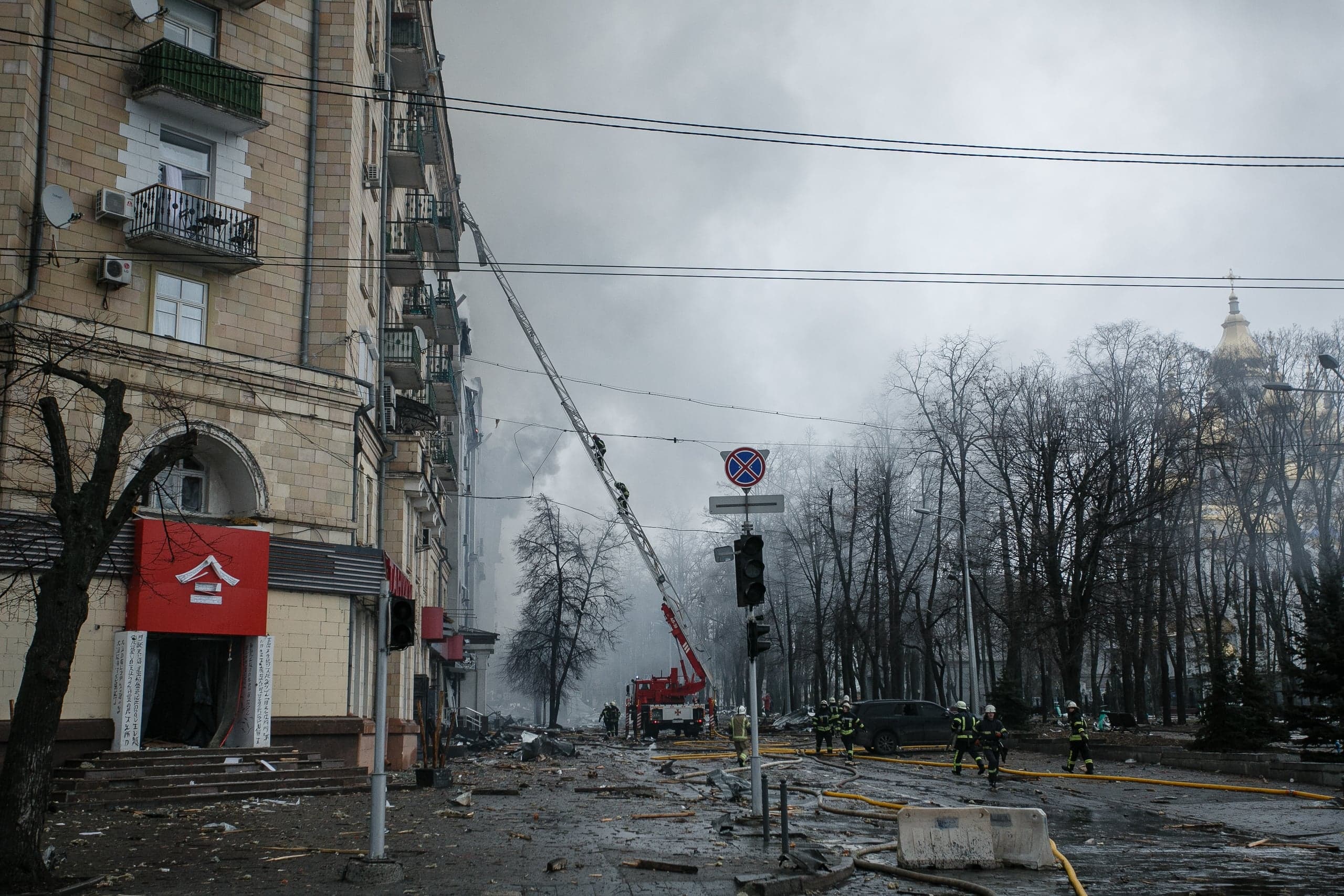 Поврежденный многоэтажный дом, из которого идет дым. Пожарные тушат огонь с помощью длинной лестницы на улице, заваленной обломками.