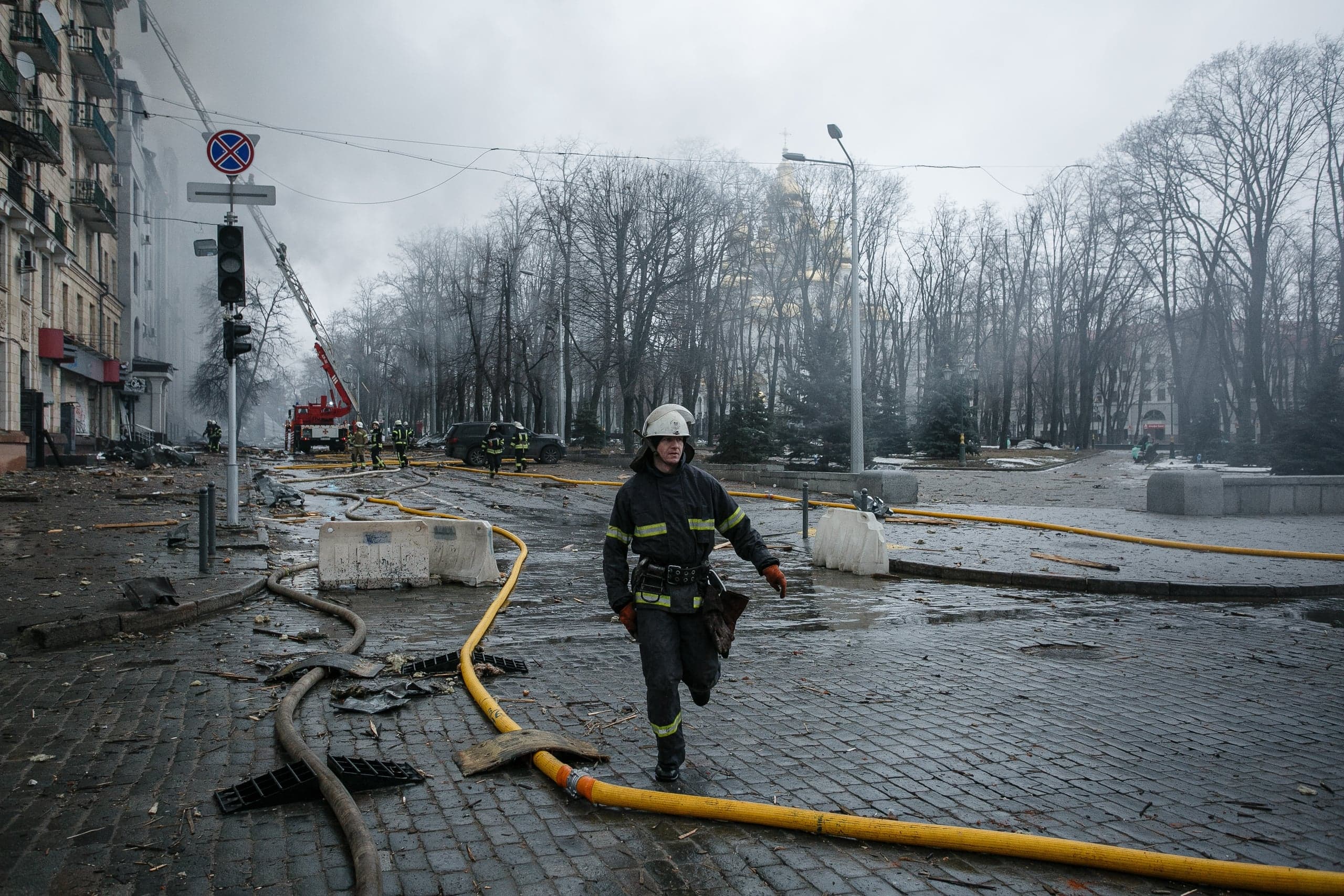 Пожарный в каске идет по разрушенной городской улице, заваленной обломками и желтыми пожарными шлангами. На заднем плане работают другие спасатели у пожарной машины с выдвижной лестницей.
