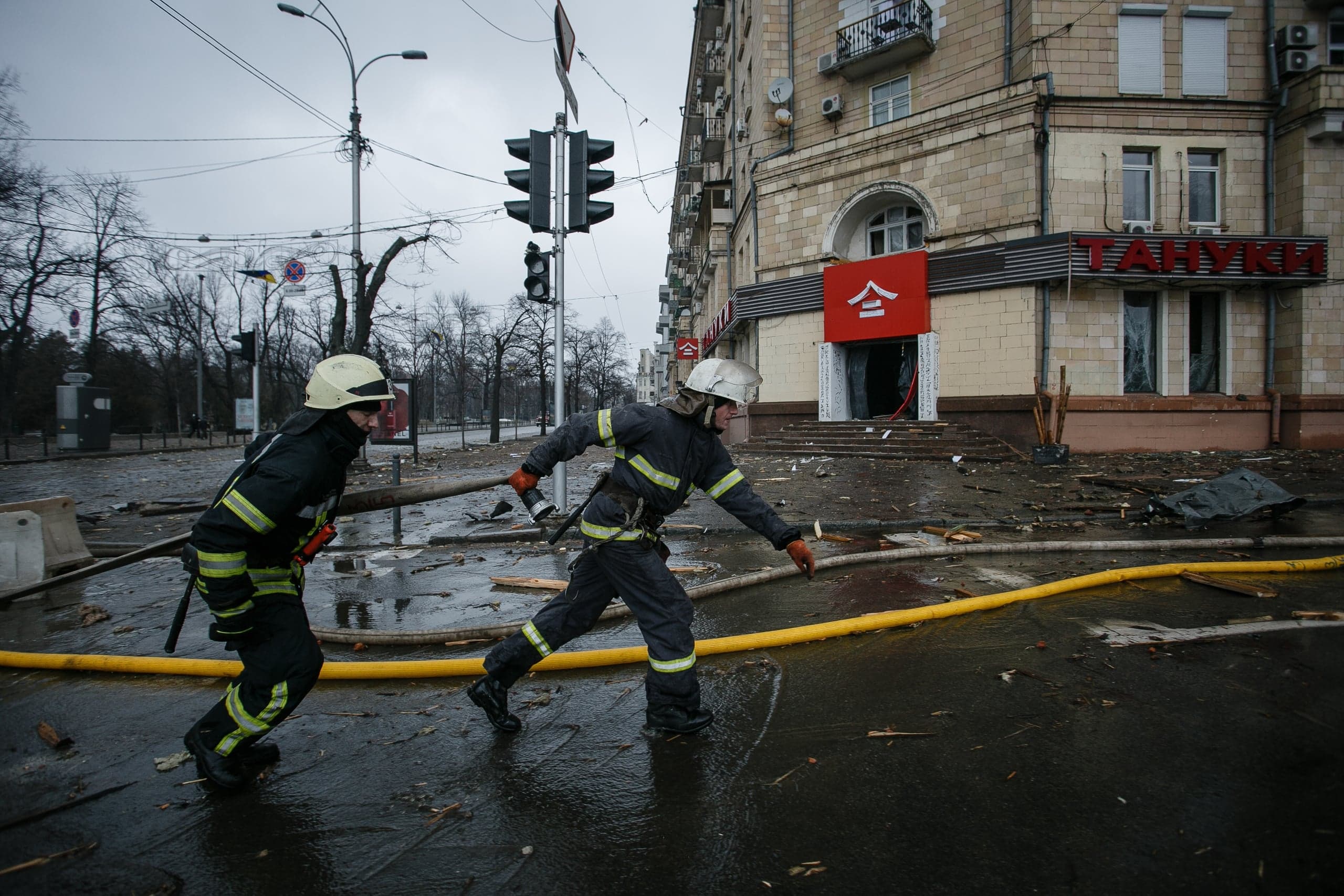 Два пожарных бегут по заваленной обломками и залитой водой улице мимо поврежденного здания с вывеской "ТАНУКИ". На земле лежит желтый пожарный рукав.
