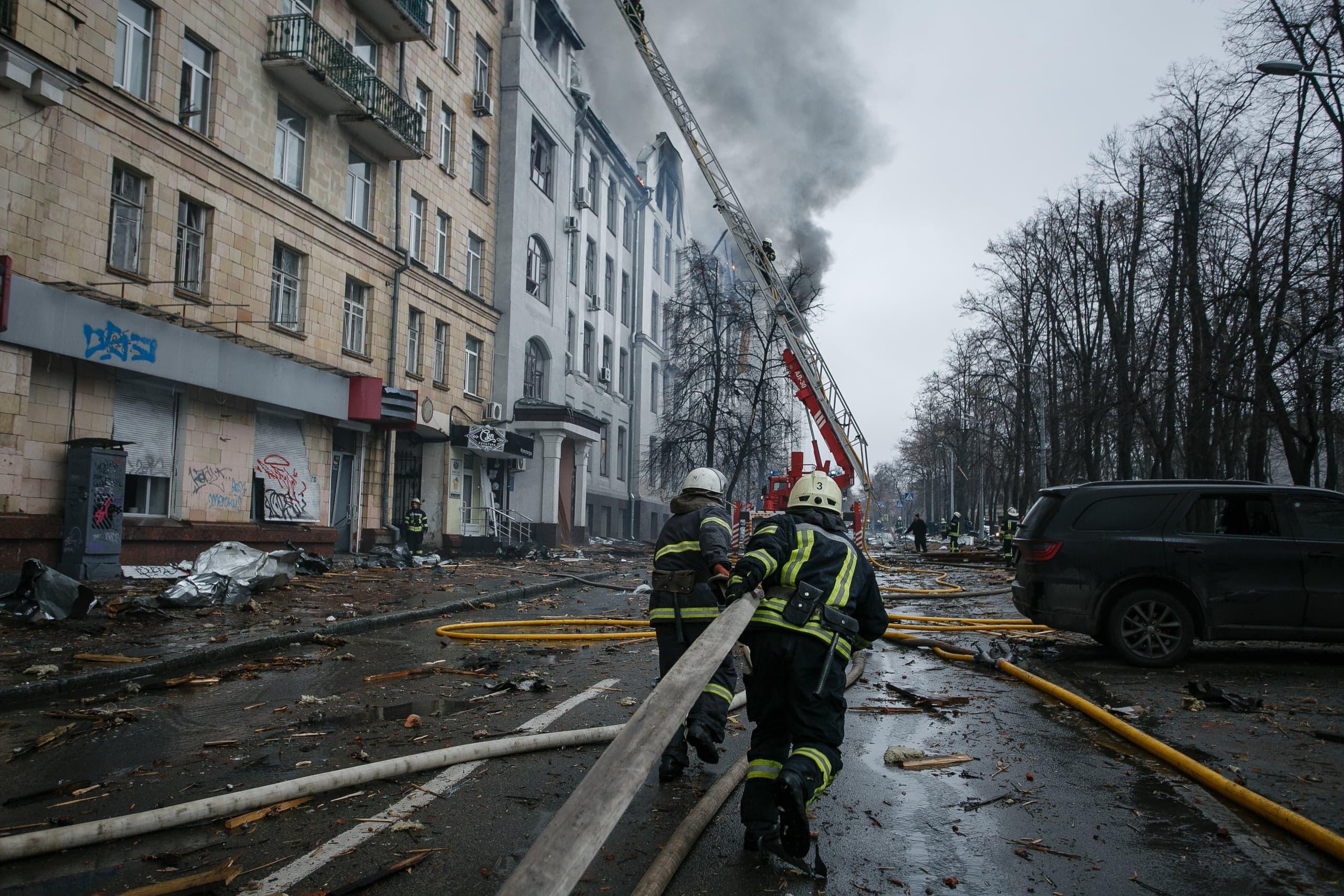 Два пожарных тянут шланг по усеянной обломками улице; из верхних этажей поврежденного здания поднимается густой дым.