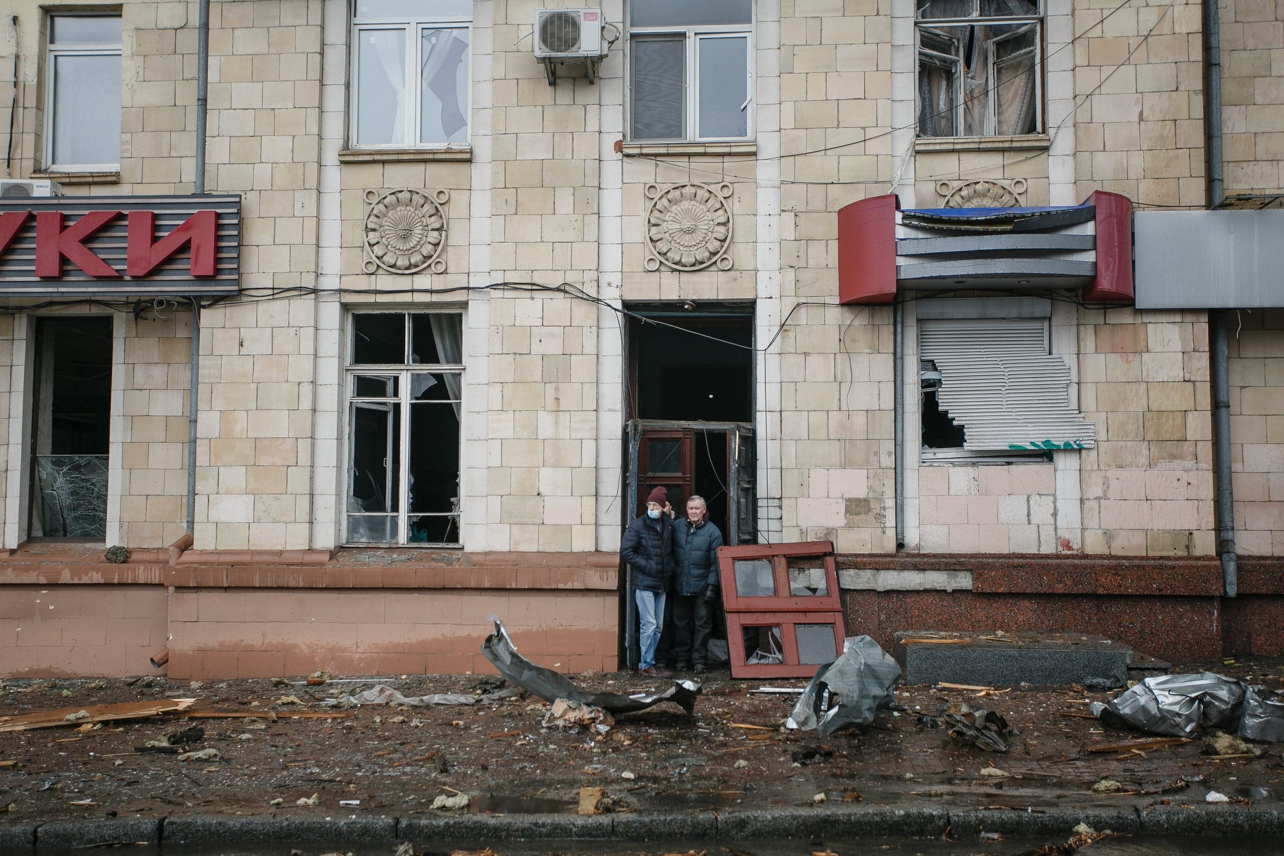 Поврежденный фасад здания с выбитыми окнами и вывеской "КИ". Двое мужчин стоят в разрушенном дверном проеме среди обломков.