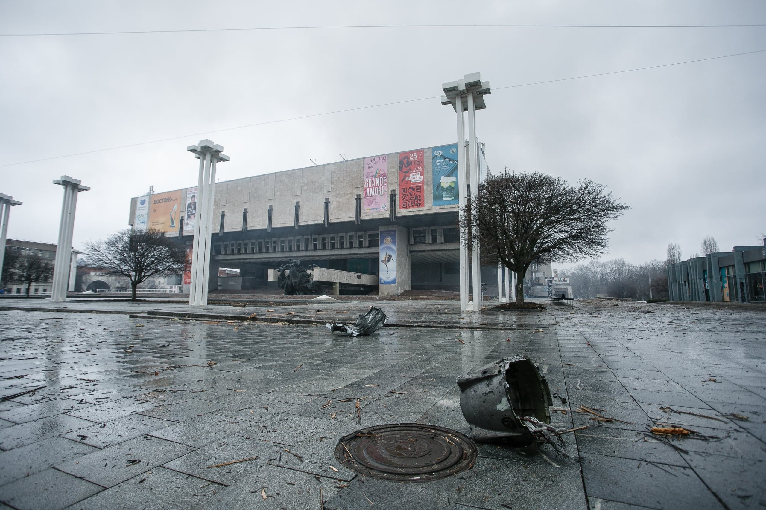 Разбитый плафон фонаря на мокрой площади перед массивным зданием культурного центра в пасмурный день.