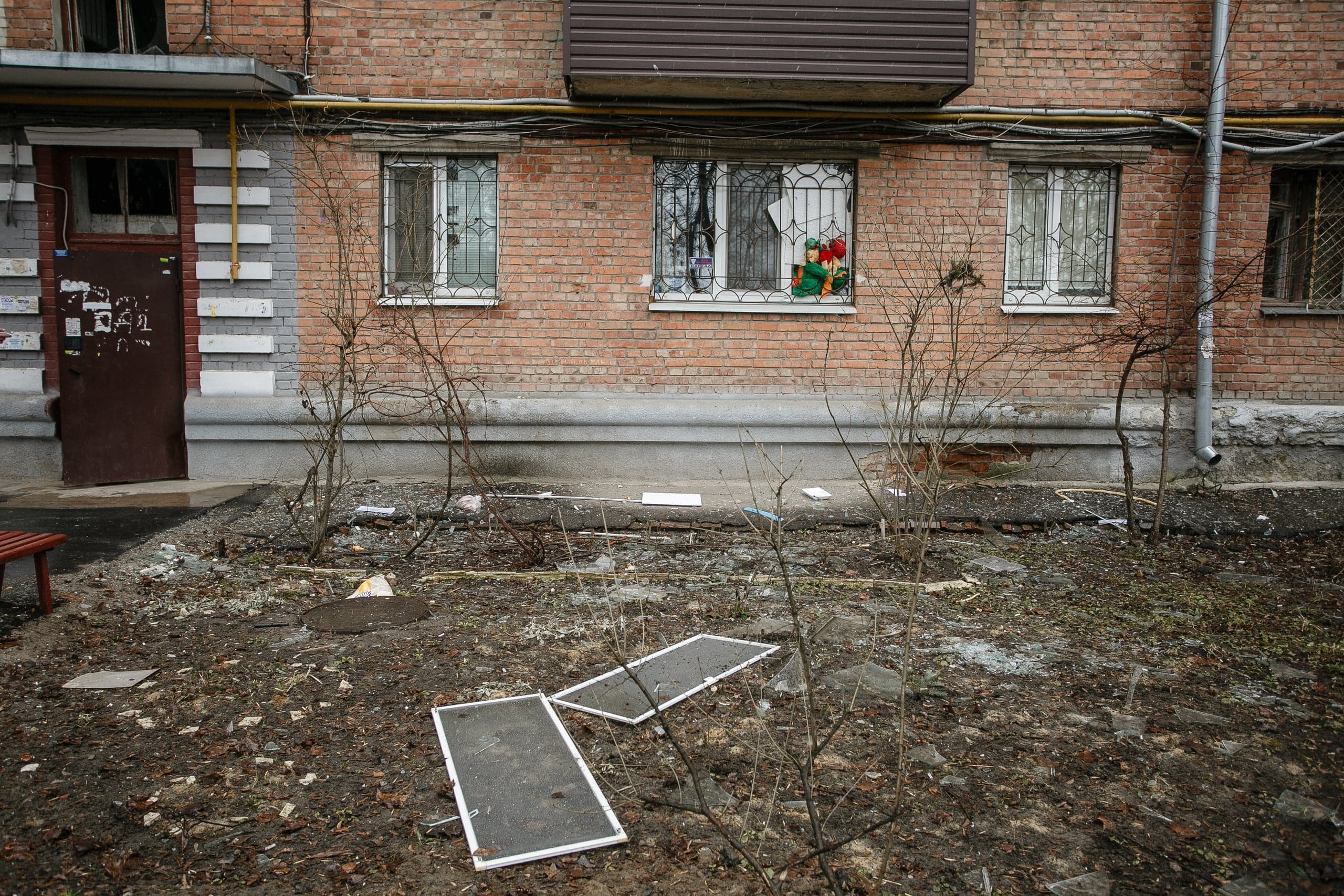 Разрушенная территория перед кирпичным жилым домом. На земле осколки стекла и оконные рамы. В разбитом окне с решеткой видны детские игрушки.