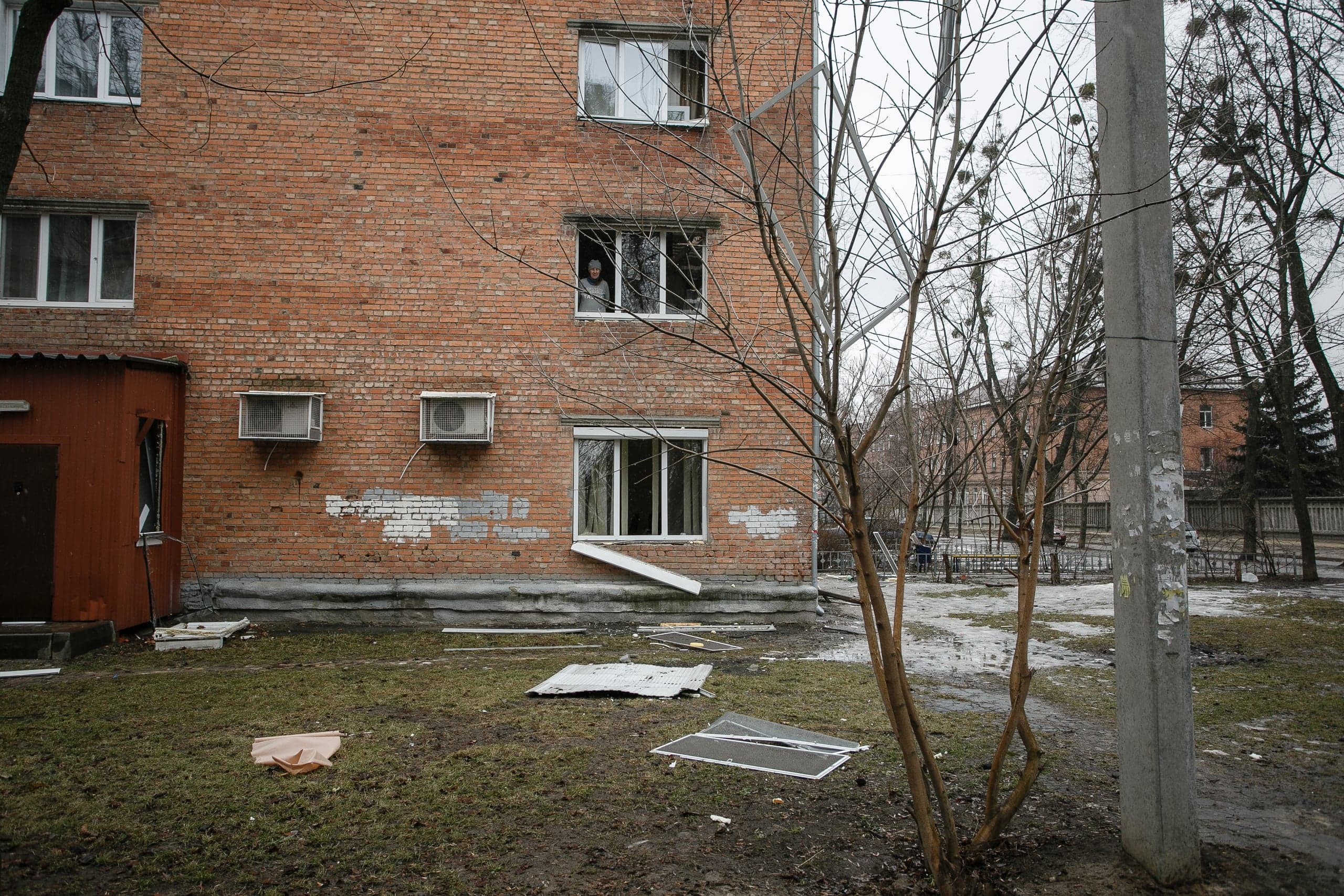 Фасад поврежденного кирпичного жилого дома; обломки окна лежат на земле. Женщина смотрит из окна на втором этаже.