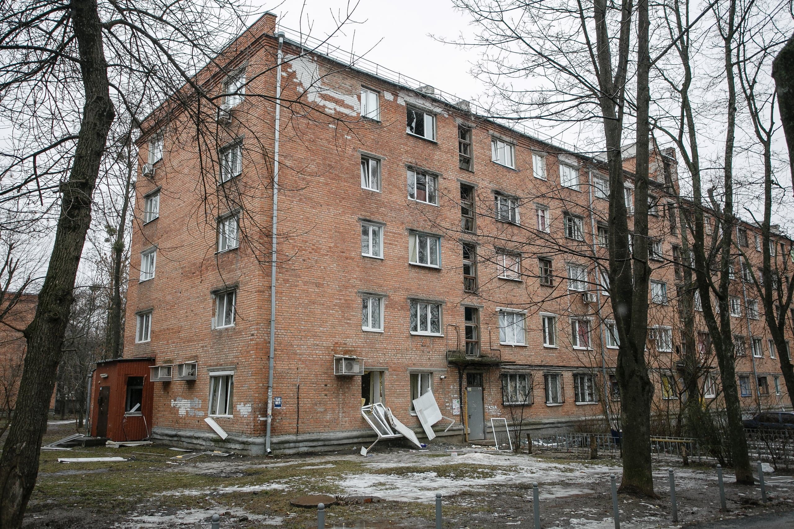 Поврежденный четырехэтажный кирпичный жилой дом, многие окна выбиты или разбиты, обломки рам лежат на земле.