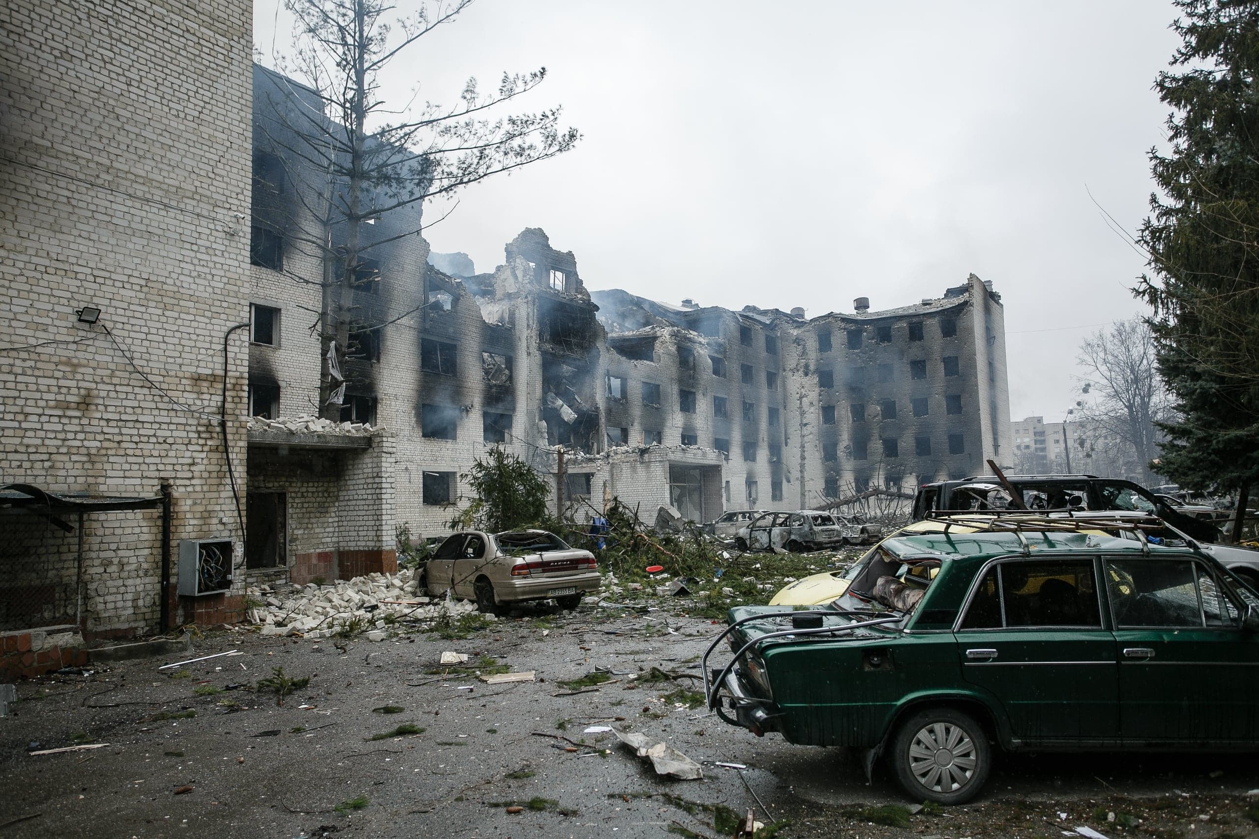 Разрушенное, обгоревшее здание общежития или жилого дома и несколько поврежденных автомобилей на засыпанной обломками площади.