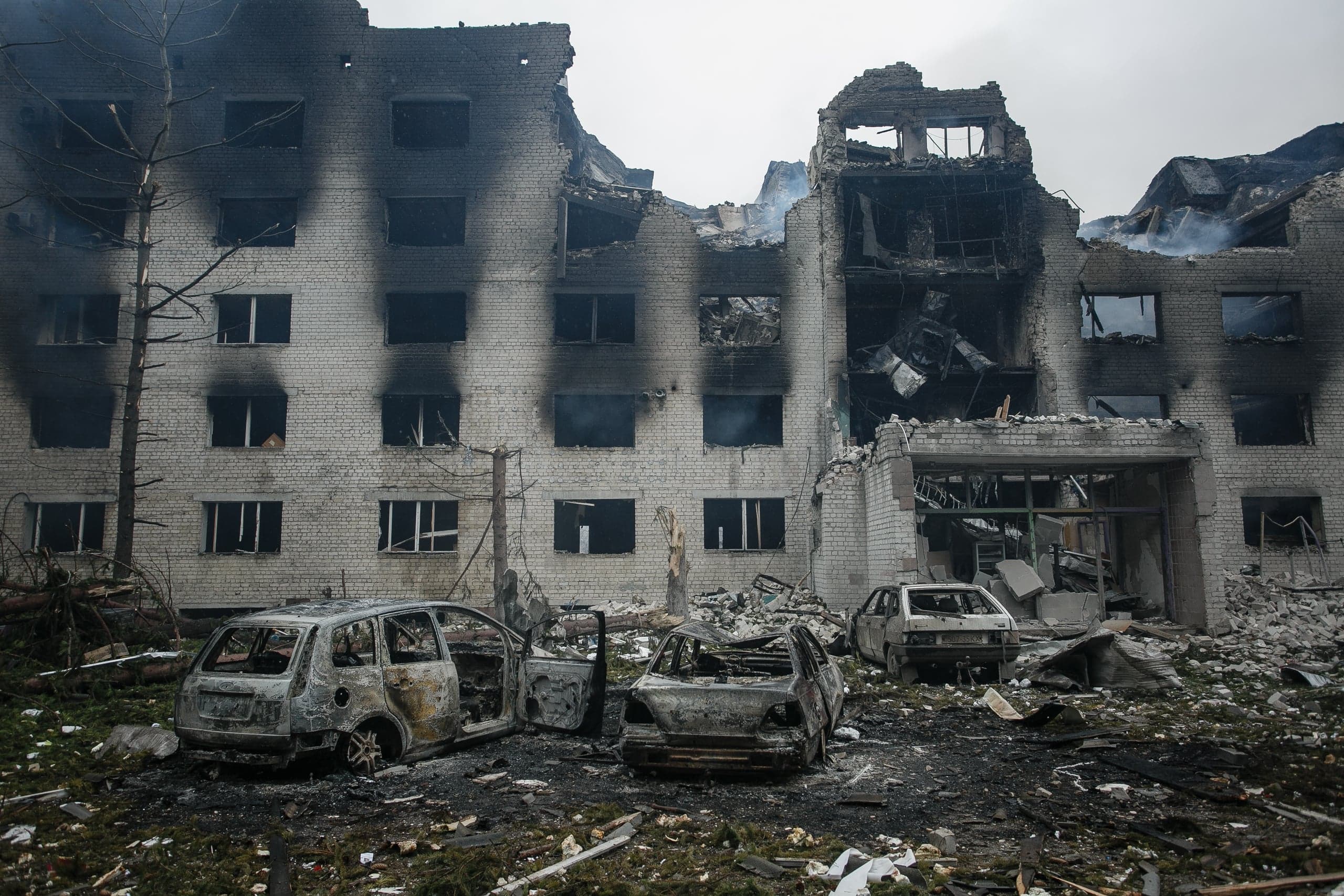 Сильно разрушенное кирпичное здание с черными следами копоти и три сгоревших автомобиля на переднем плане.