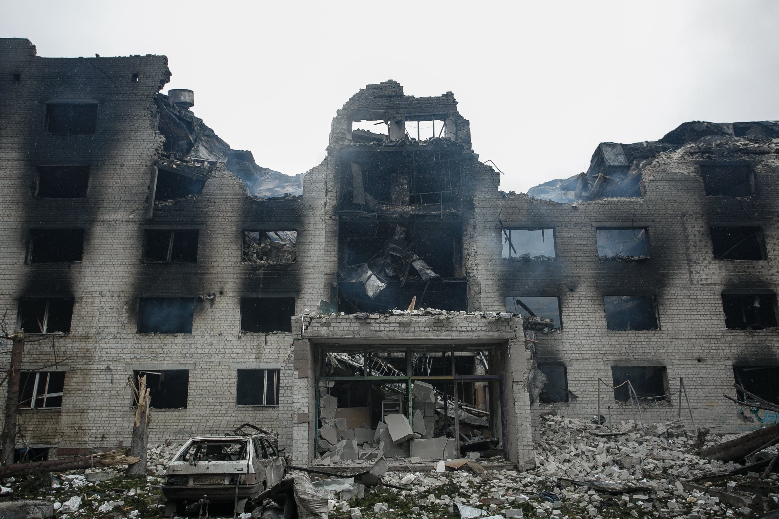 Разрушенное взрывом кирпичное здание: черный от копоти фасад, обрушенная центральная часть, груды обломков и уничтоженный автомобиль.