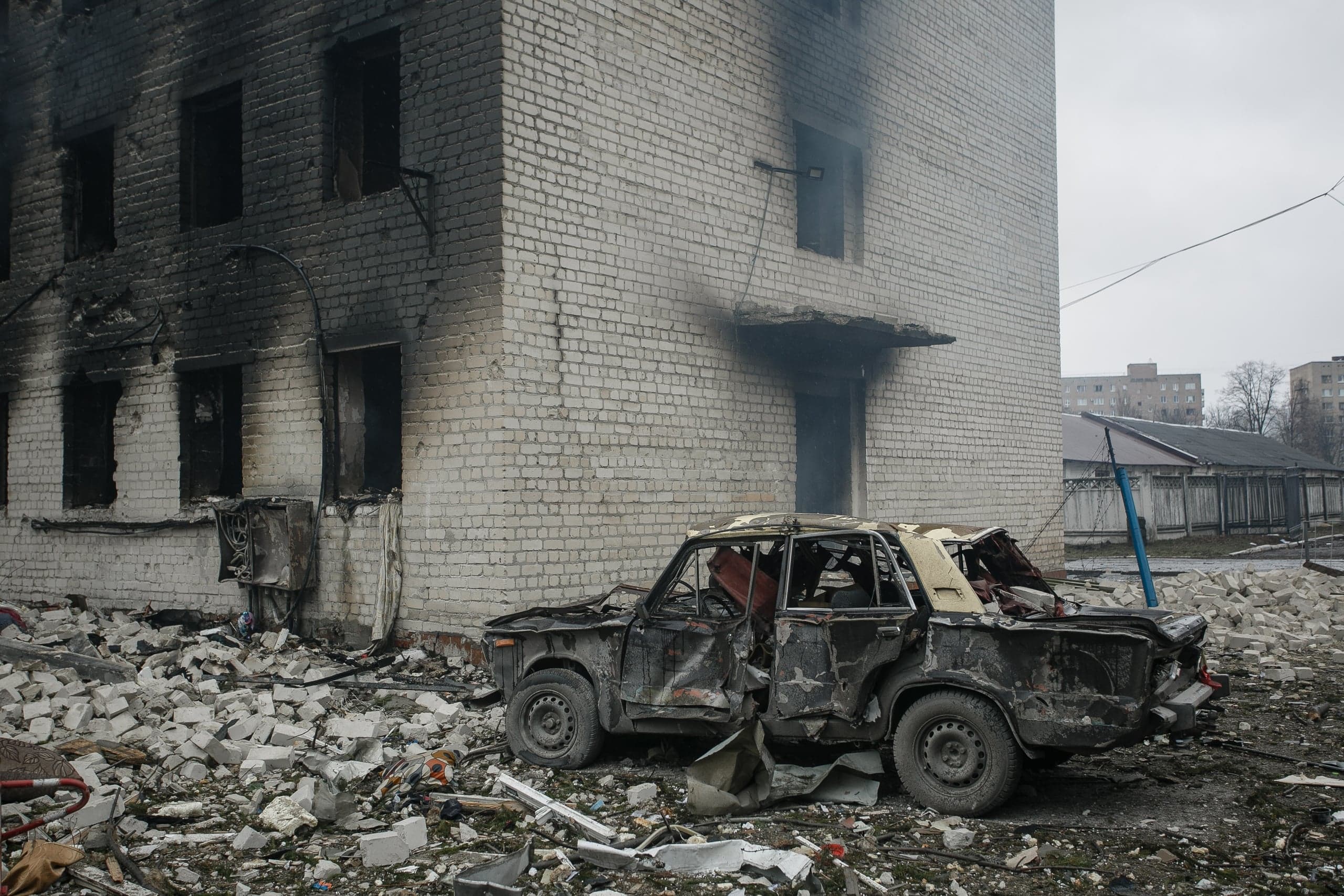 Сгоревший автомобиль на фоне закопченного обстрелом кирпичного здания, окруженного грудами обломков.