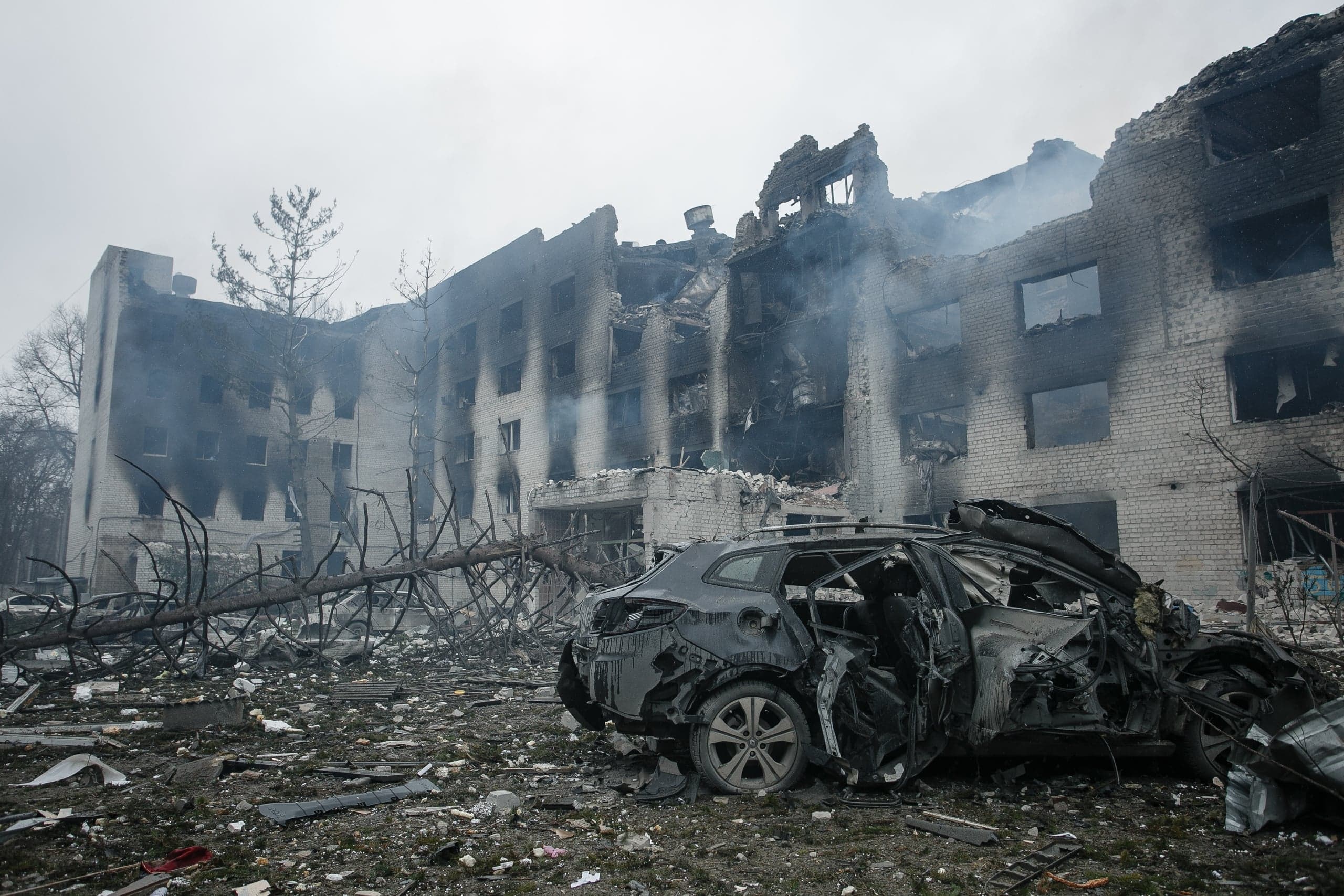 Разрушенное обгоревшее здание, из которого идет дым. На переднем плане — искореженный автомобиль среди обломков.