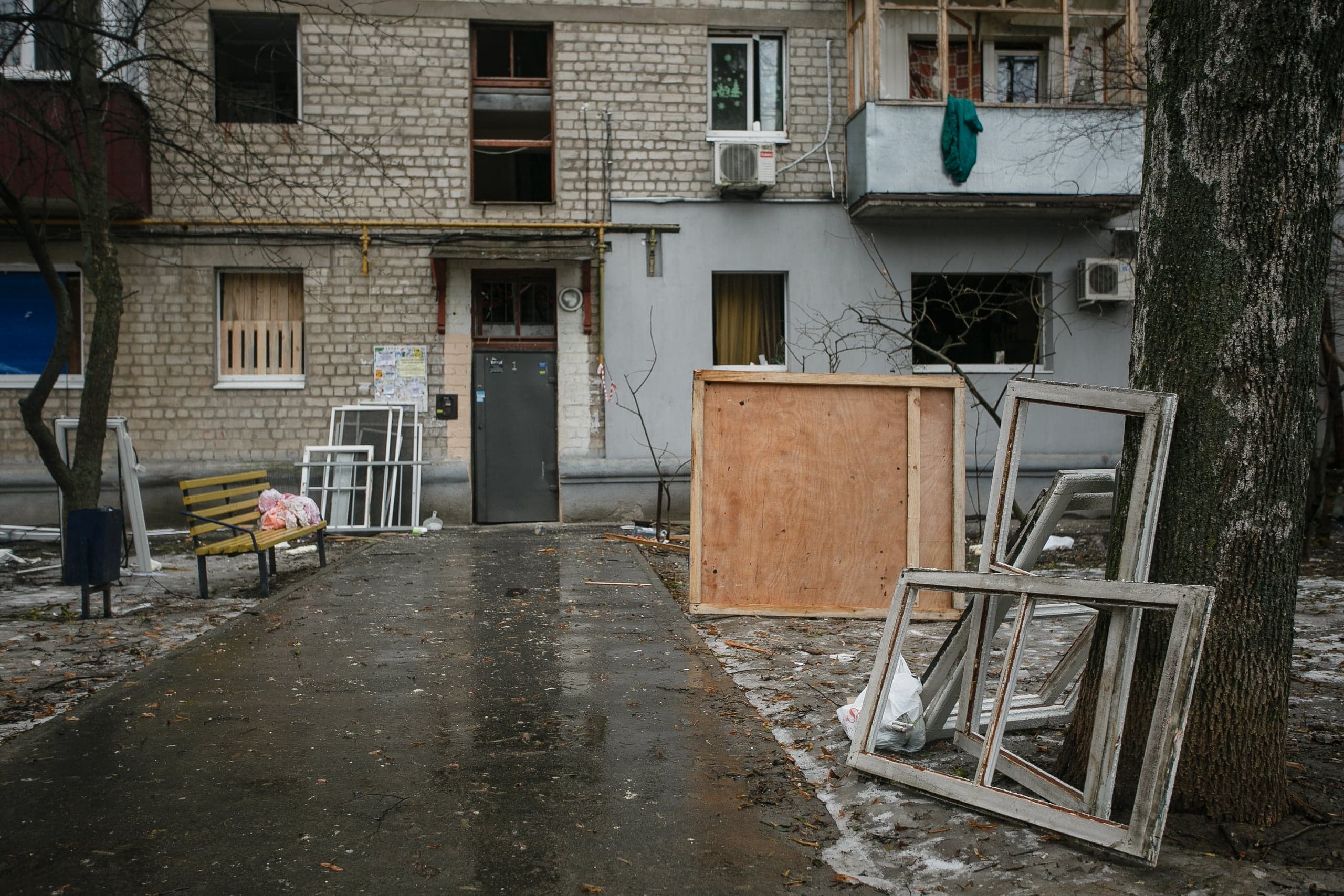 Подъезд поврежденного кирпичного дома с выбитыми окнами. У входа сложены новые оконные рамы для замены; старые рамы лежат у дерева на мокром тротуаре.