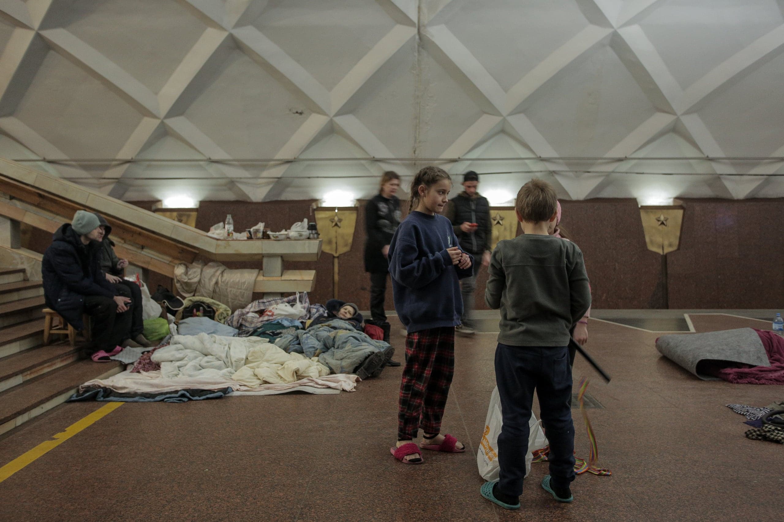 Люди, включая детей, укрываются на платформе метро: спят на полу, укрывшись одеялами, и стоят в ожидании.