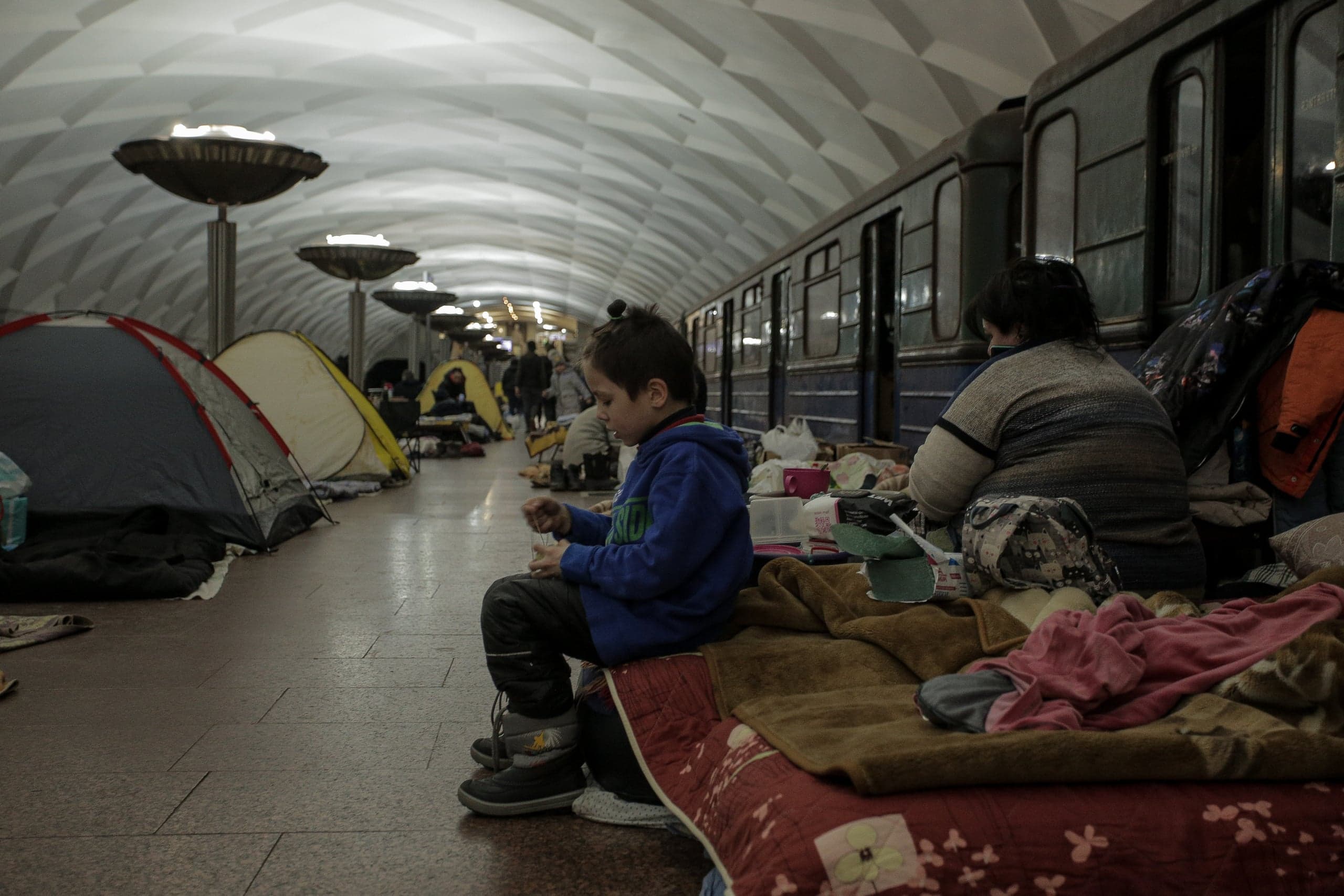 Станция метро, используемая как убежище. Палатки и матрасы расположены вдоль платформы, у вагона поезда сидят ребенок и женщина.