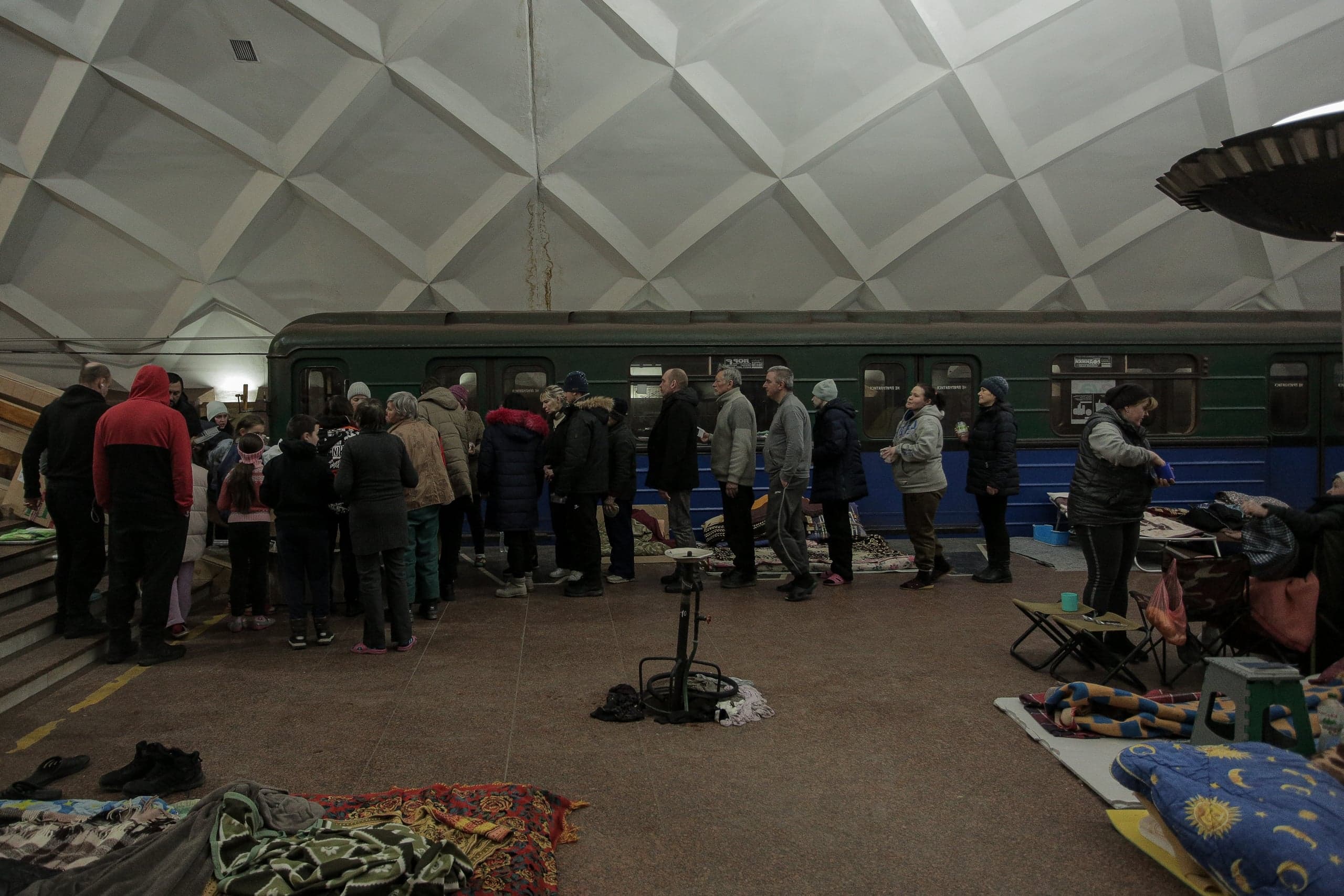 Люди укрываются на платформе метро, стоя вдоль неподвижного поезда. Пол станции покрыт матрасами, одеялами и личными вещами.