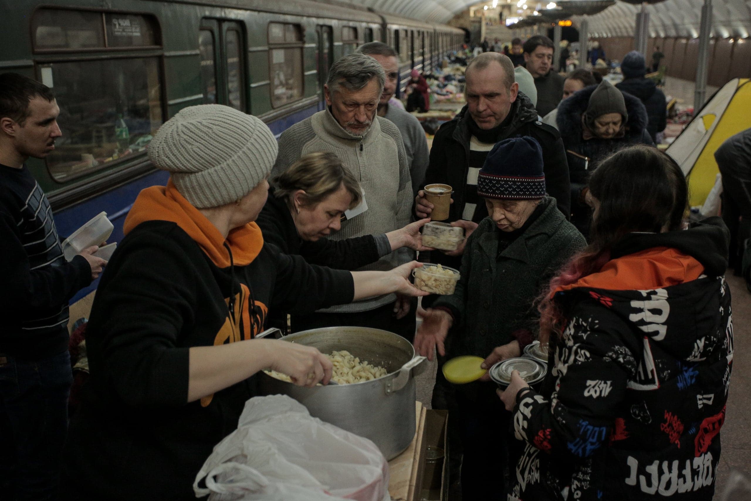 Волонтер раздает еду (пасту) из большого котла людям, ищущим убежище на платформе метро. На заднем плане виден припаркованный поезд.