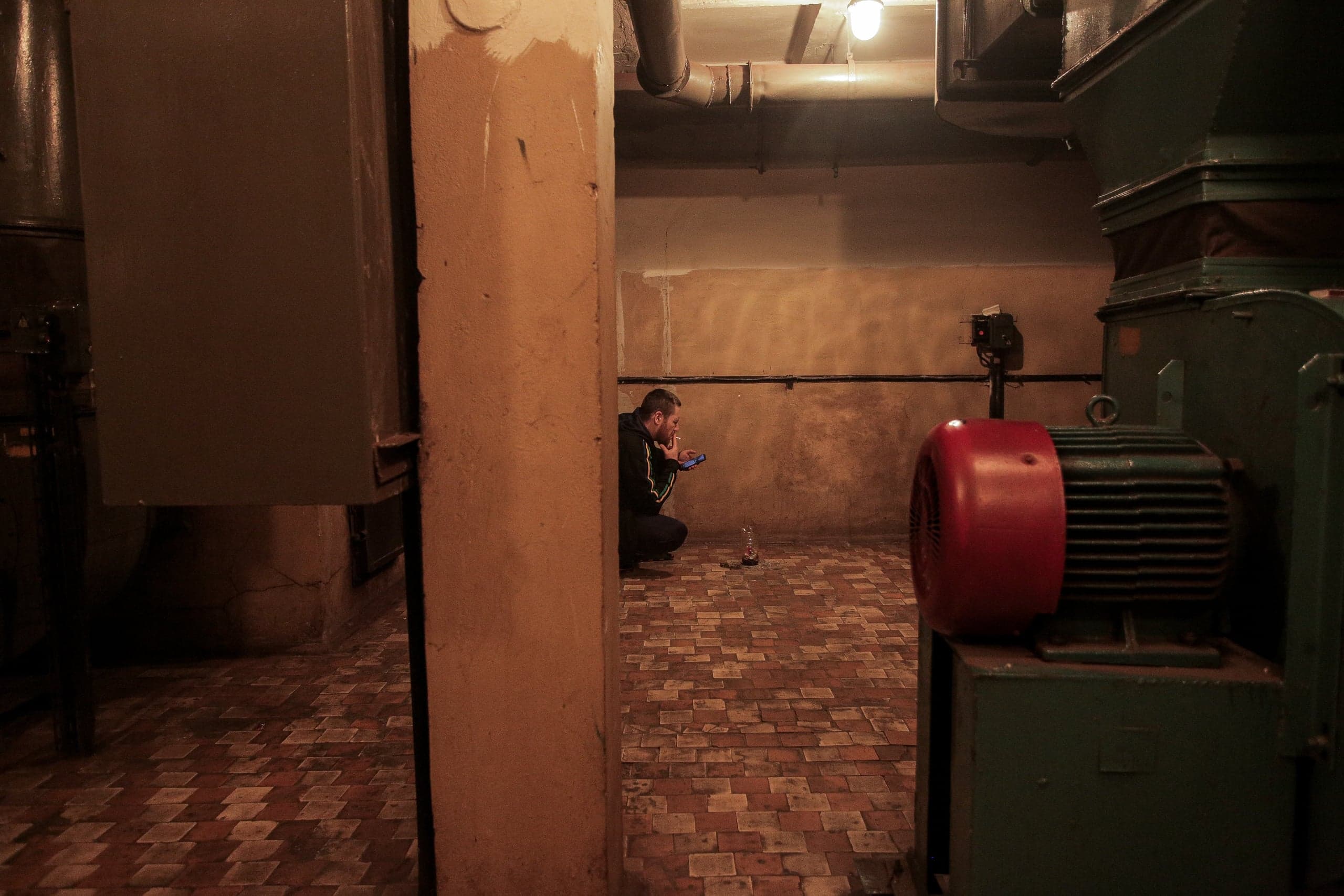Мужчина сидит на корточках в полутемном подвальном помещении среди труб и оборудования. Он курит и смотрит в телефон; справа — крупный агрегат с красным двигателем.