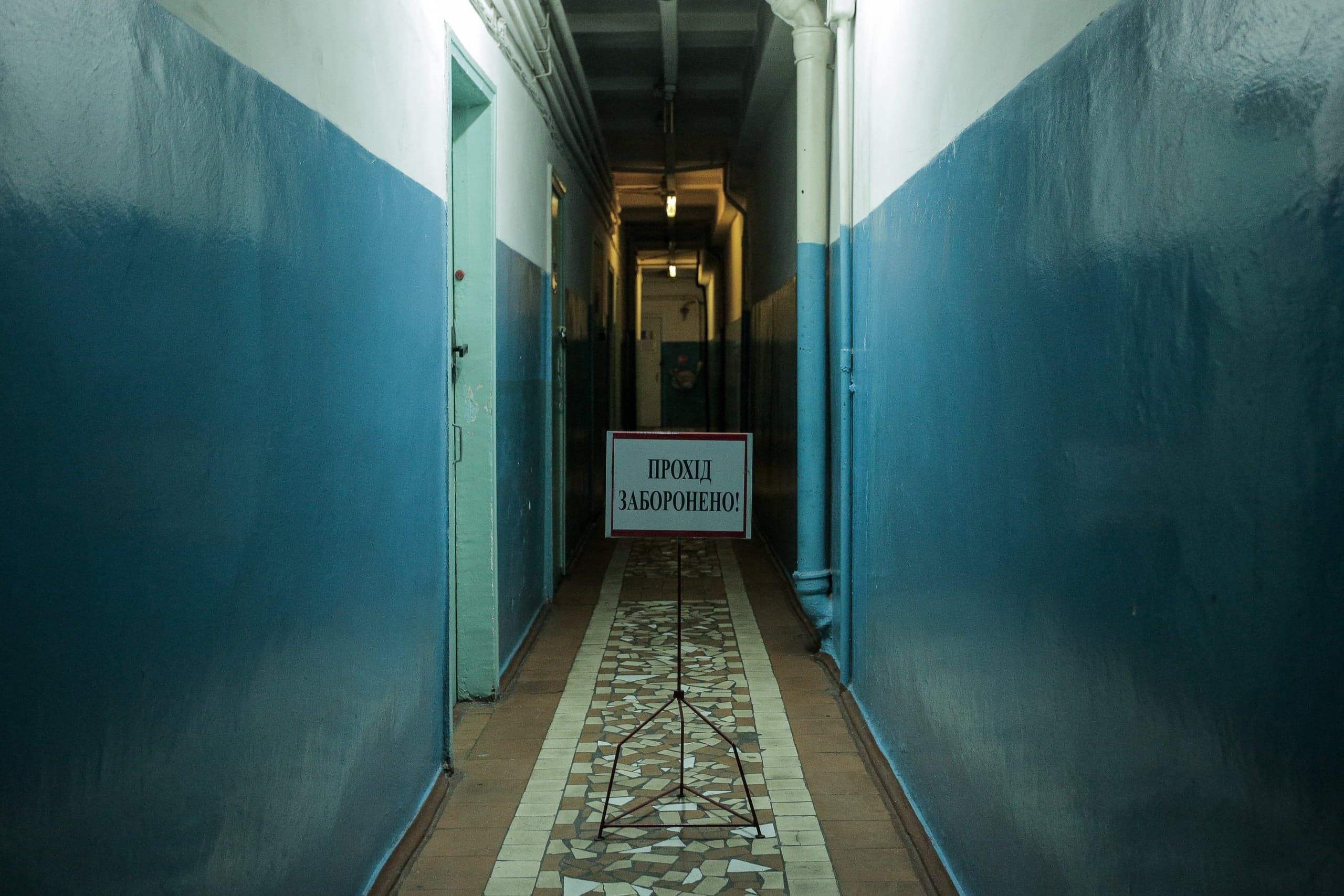 Узкий коридор с сине-белыми стенами и мозаичным полом. В центре проход перекрыт знаком: "ПРОХІД ЗАБОРОНЕНО!".