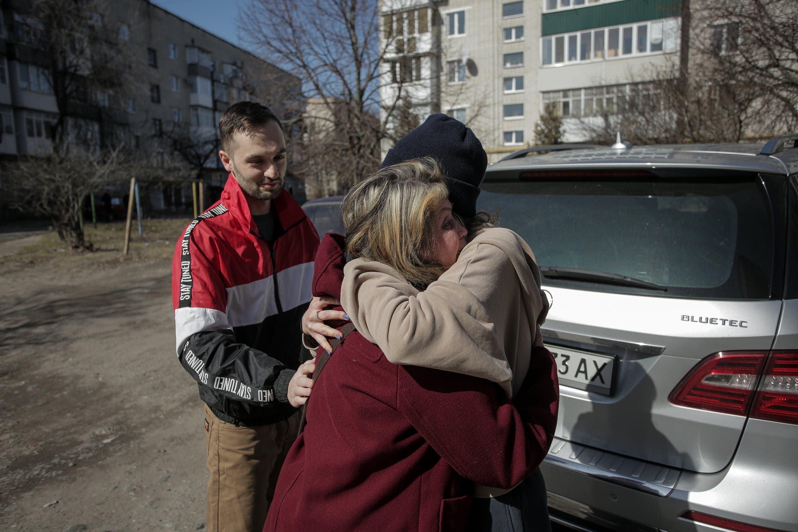 Женщина и человек в капюшоне обнимаются; сбоку стоит мужчина в куртке с надписью "STAY TUNED". Фон: жилые дома и серебристый автомобиль с маркировкой "BLUETEC".