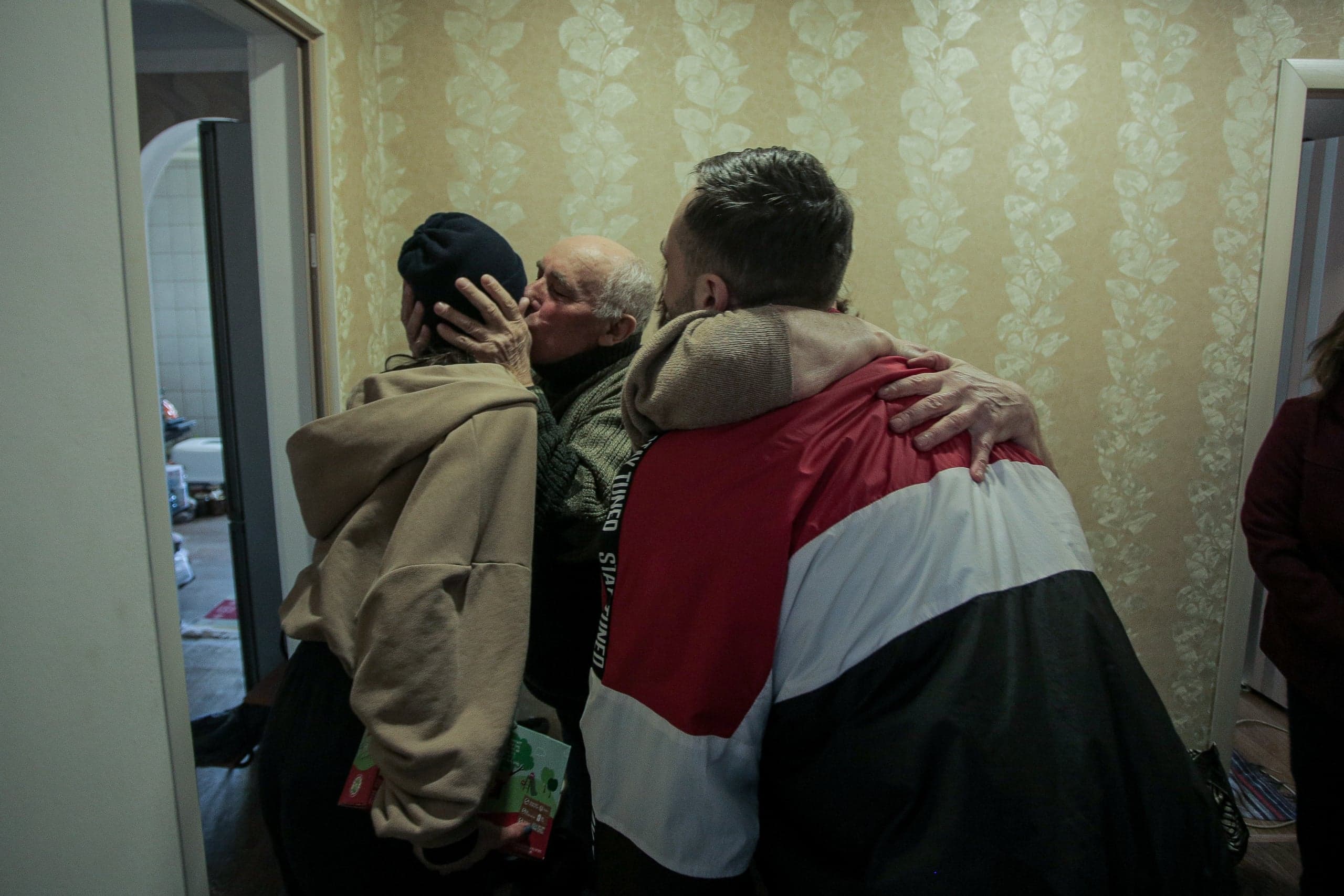 Пожилой мужчина целует женщину в черной шапке, обнимая при этом другого мужчину в красно-бело-черной куртке, на фоне обоев с цветочным узором.