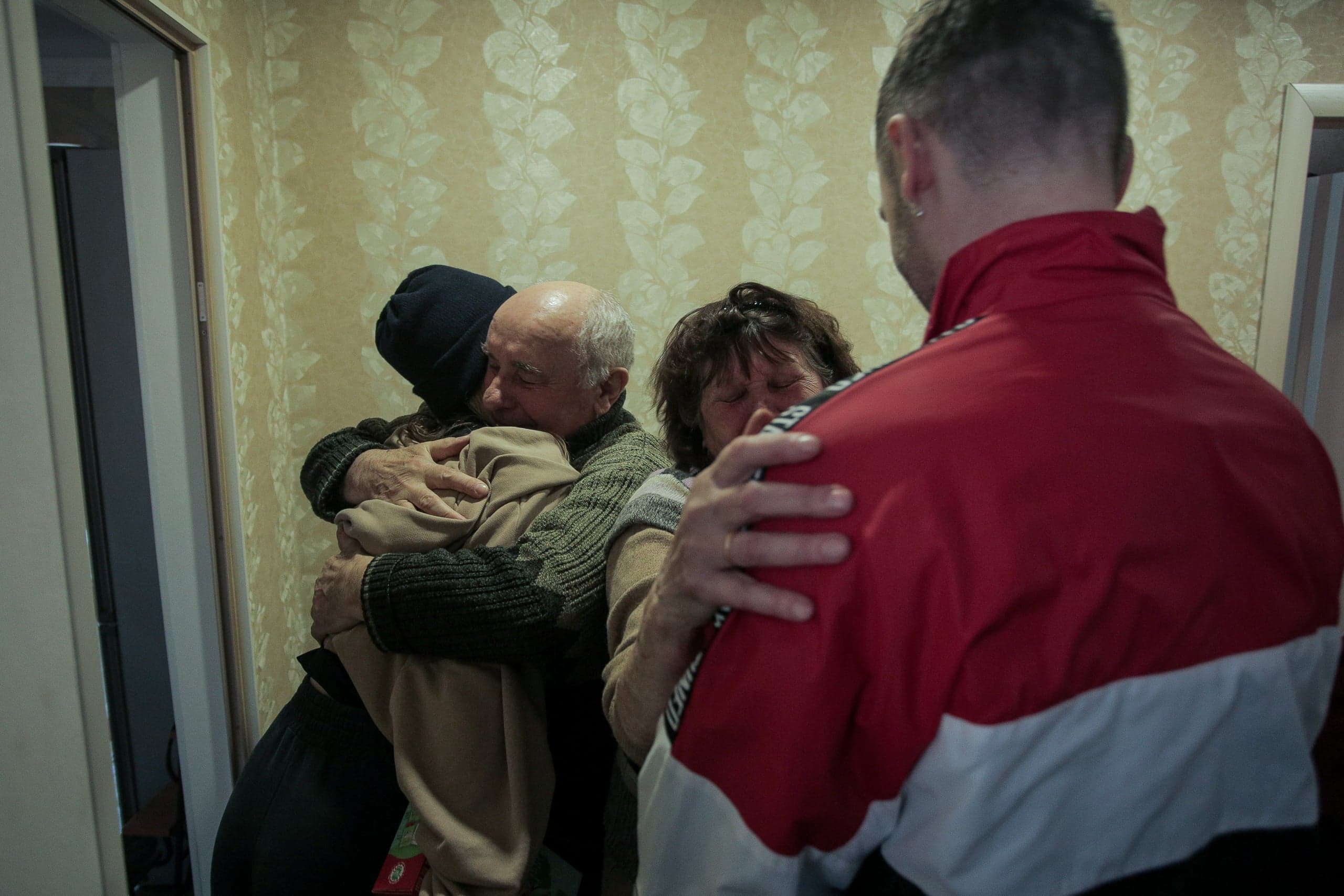 Четыре человека в эмоциональном объятии: пожилая пара обнимает двух людей в комнате с узорчатыми обоями. Справа — спина человека в красно-белой куртке.